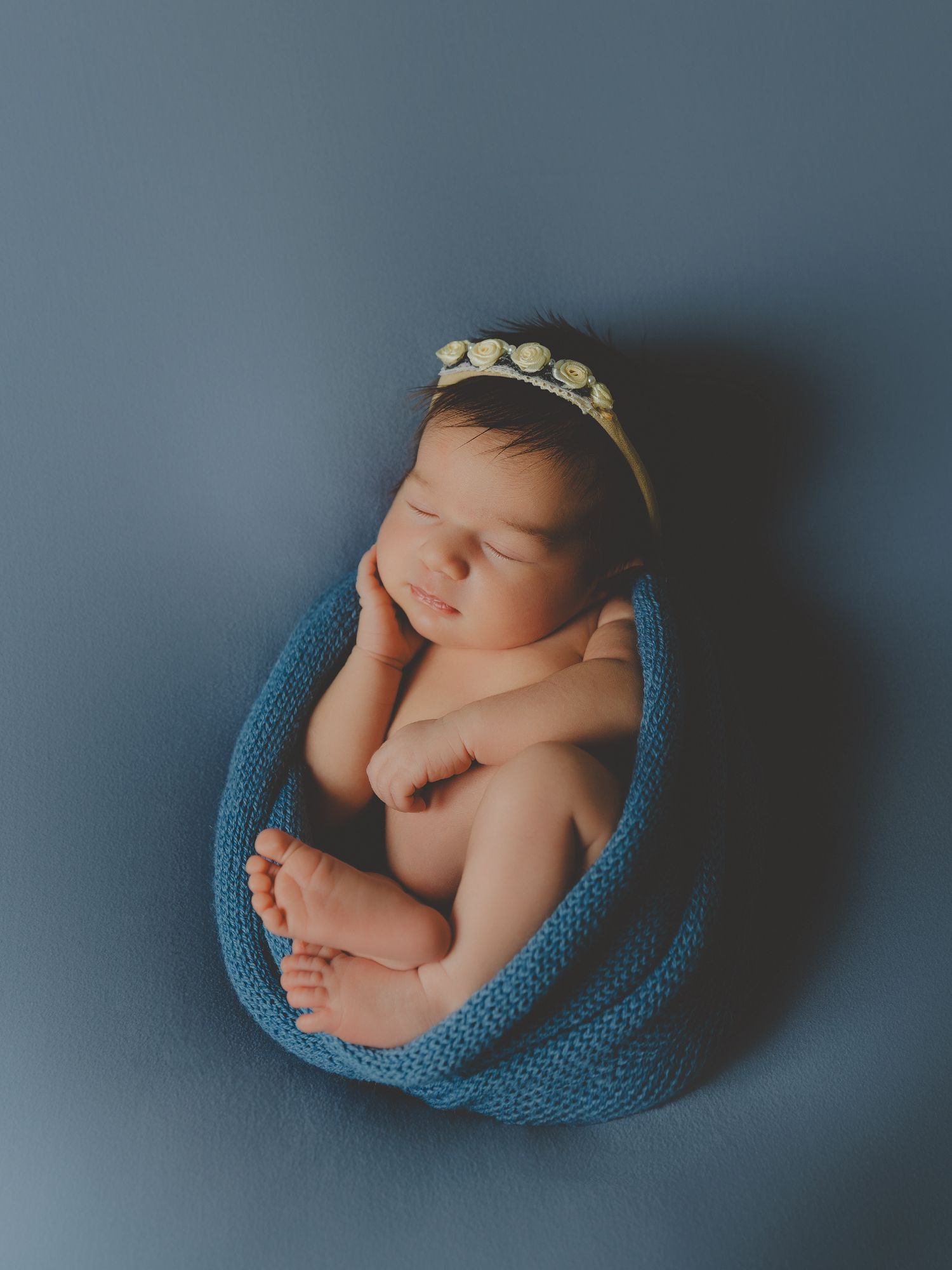 Bebé recién nacido durmiendo en su sesión, en el estudio fotográfico, envuelto en una manta de punto azul con una diadema floral amarilla.