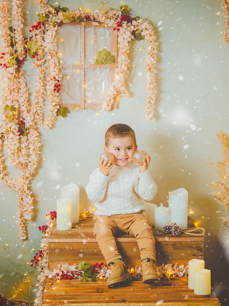 Fotografía de un niño sentado en una escalera de madera jugando con bolas de Navidad en un decorado con velas.