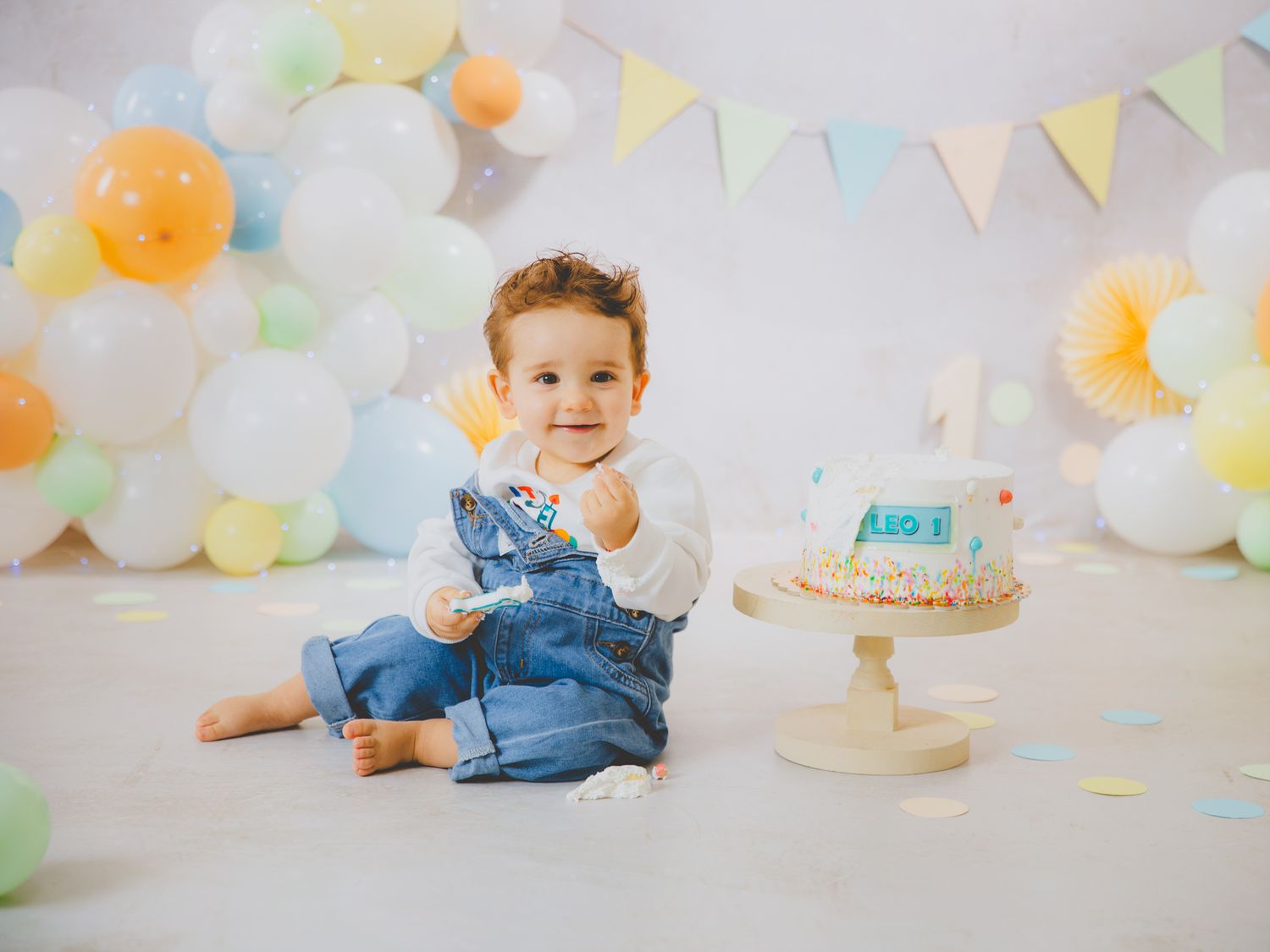Un pequeño bebé sonriente, vestido con un mono vaquero, celebra su primer cumpleaños con pastel y decoraciones de globos en tonos pastel.