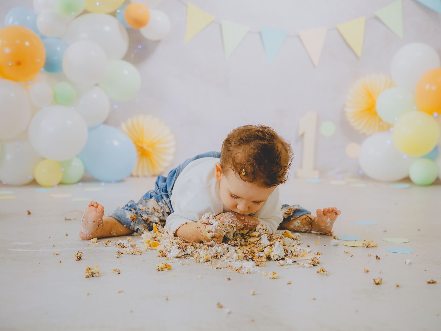 Un bebé con un peto vaquero come un pastel de cumpleaños aplastado en el suelo, con un fondo de globos.