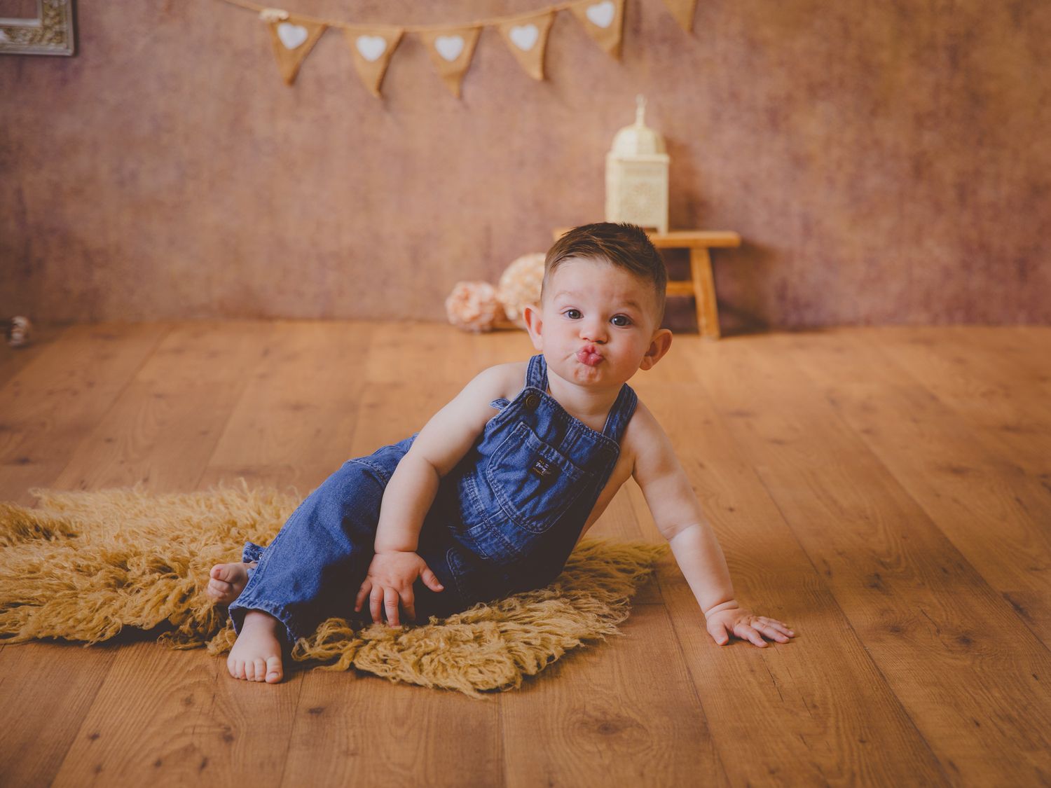 Un adorable bebé con un mono vaquero está sentado sobre una alfombra haciendo una mueca graciosa en su sesión de estudio de estilo rústico.