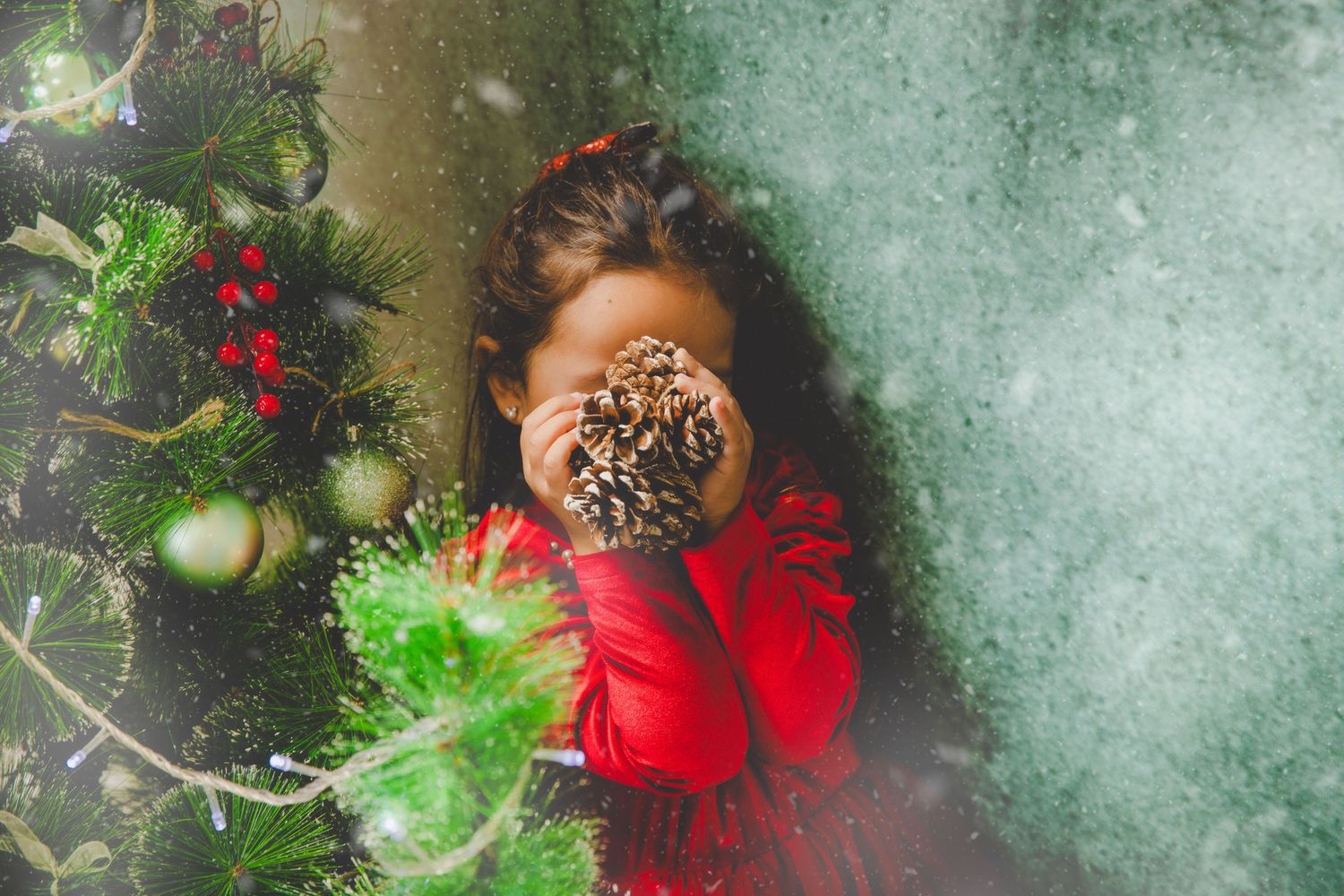 Una niña con conjunto rojo esconde su cara detrás de una piña de pino junto a un árbol de Navidad.