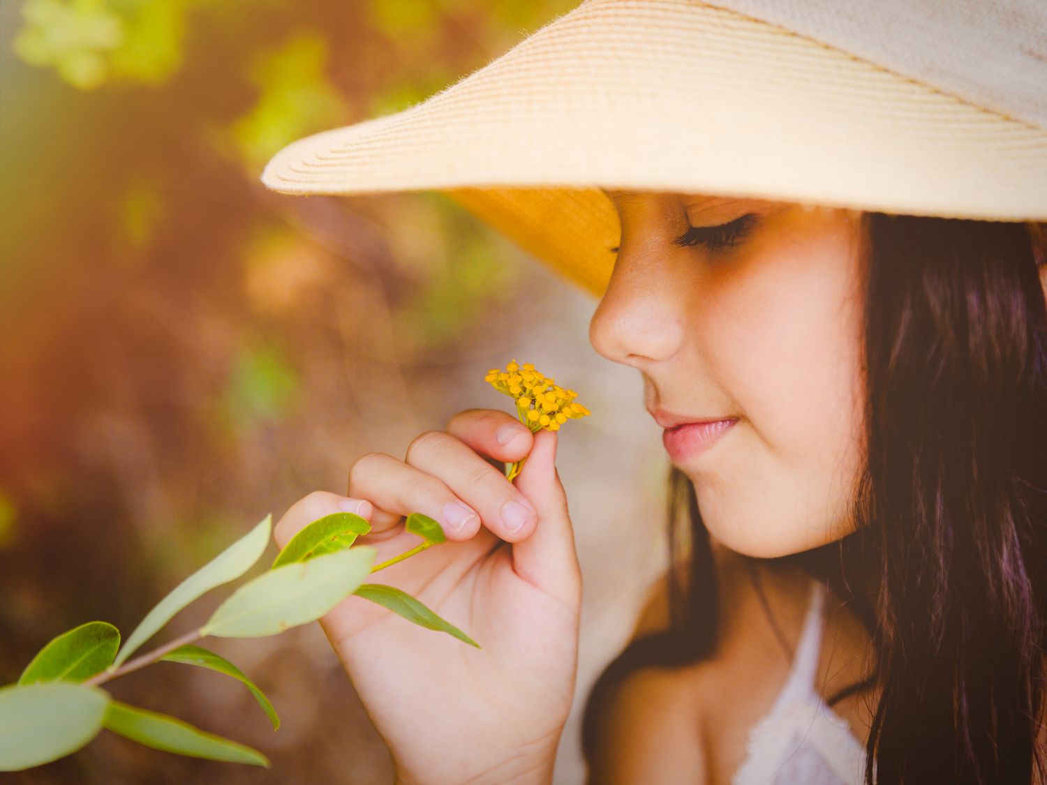 Niña con sombrero de ala ancha oliendo una flor silvestre amarilla al aire libre.