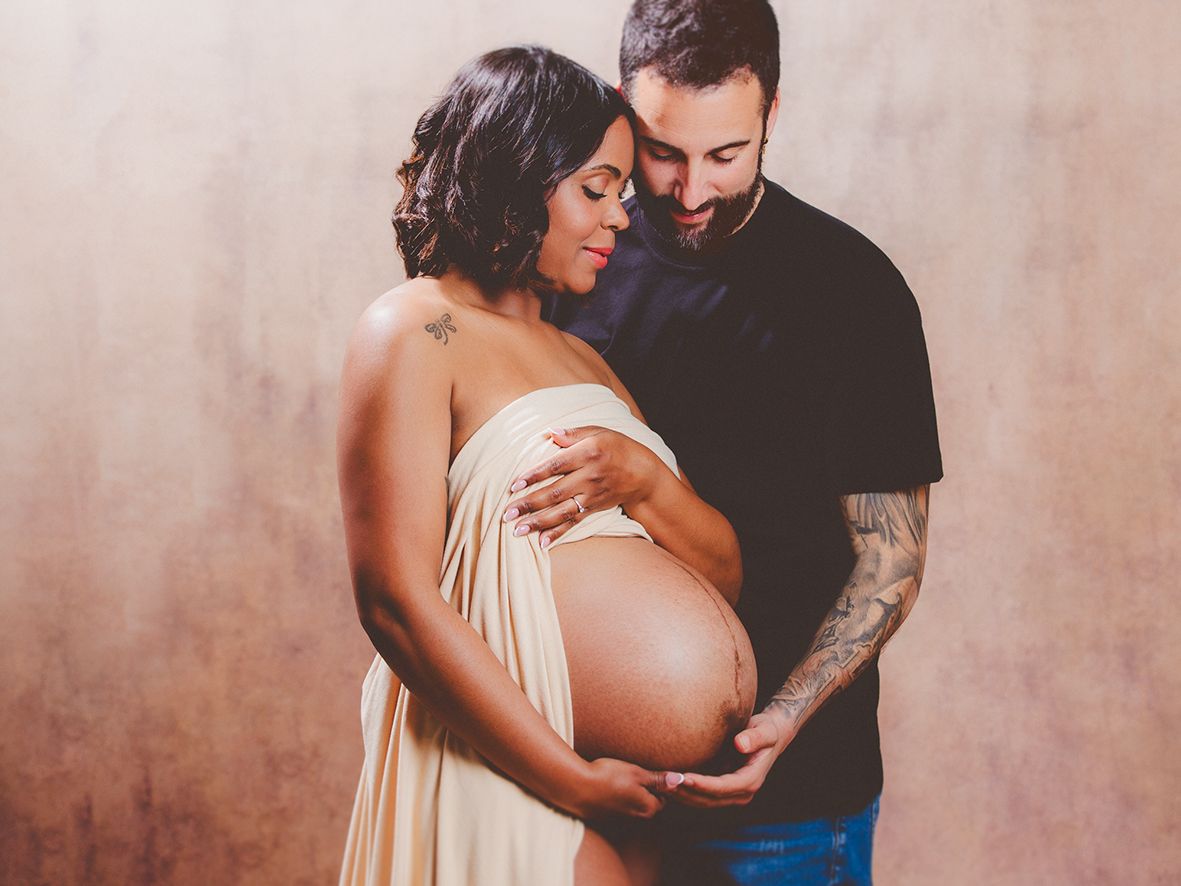 Una pareja abraza tiernamente la barriga de la futura mamá en una sesión de fotos de maternidad en estudio con tonos cálidos.