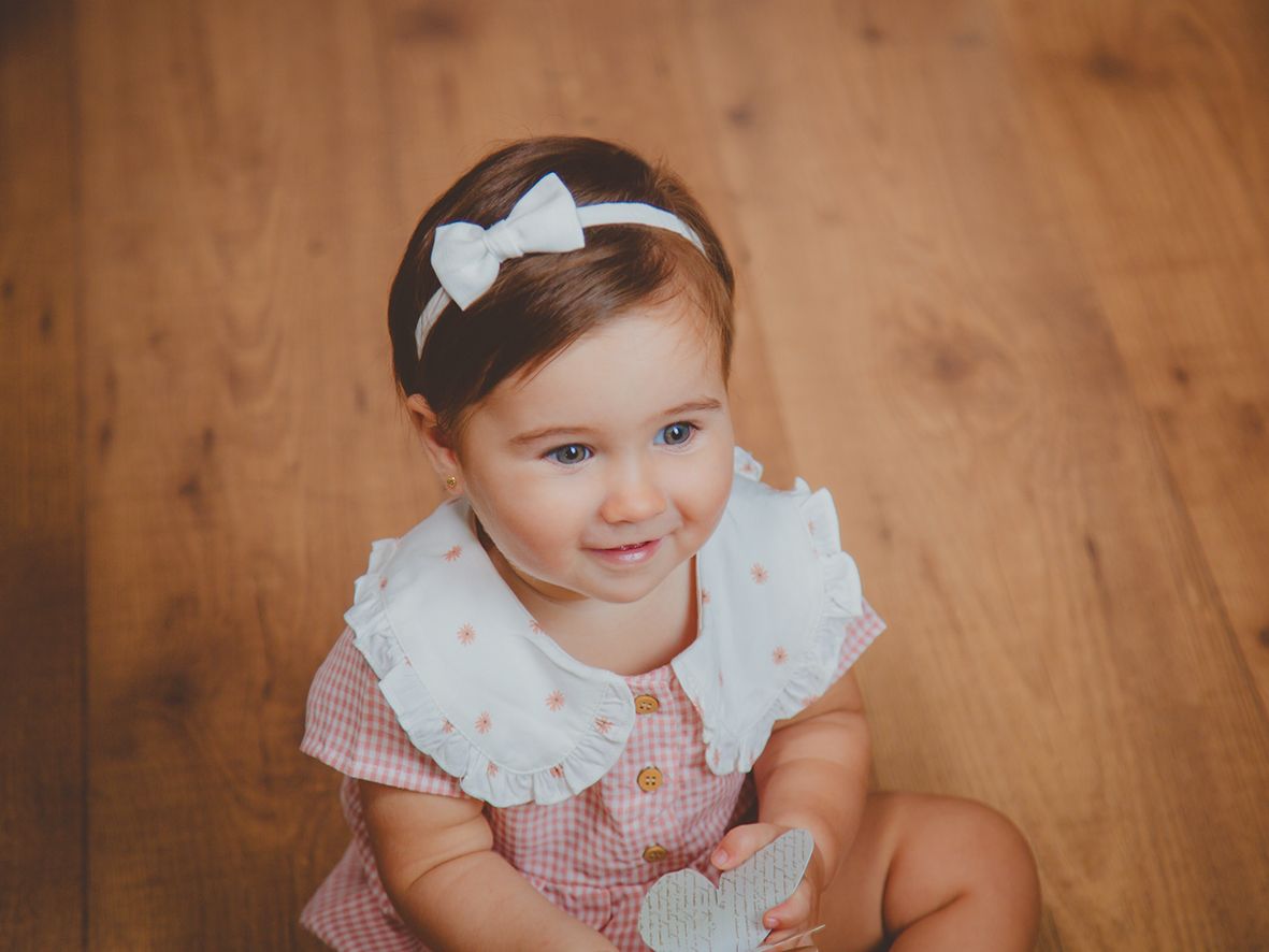 Una linda niña con un vestido rosa y diadema blanca con lazo está sentada en un suelo de madera sonriendo en su sesión de fotos de bebé.