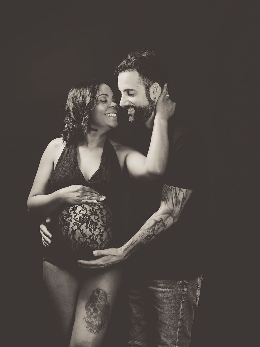 Fotografía de maternidad en blanco y negro de una pareja sonriente con tatuajes en un estudio.