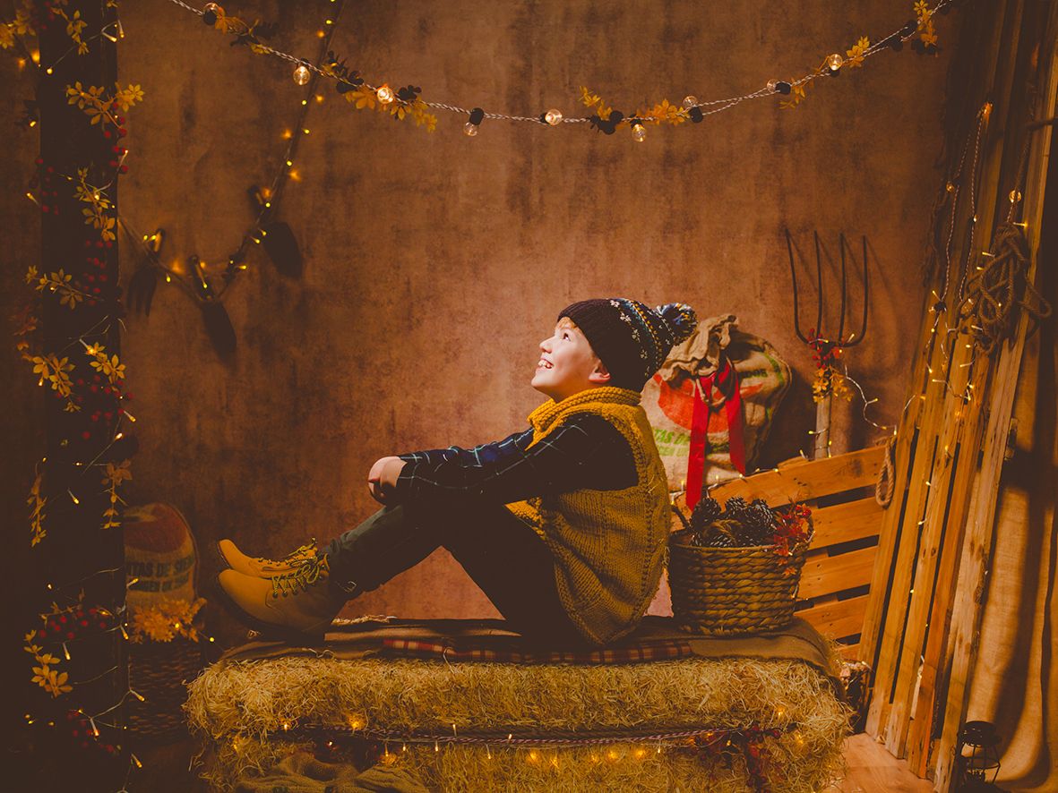 Un niño con gorro y chaleco descansa sobre fardos de heno rodeado de cálidas luces en una fotografía navideña rústica.