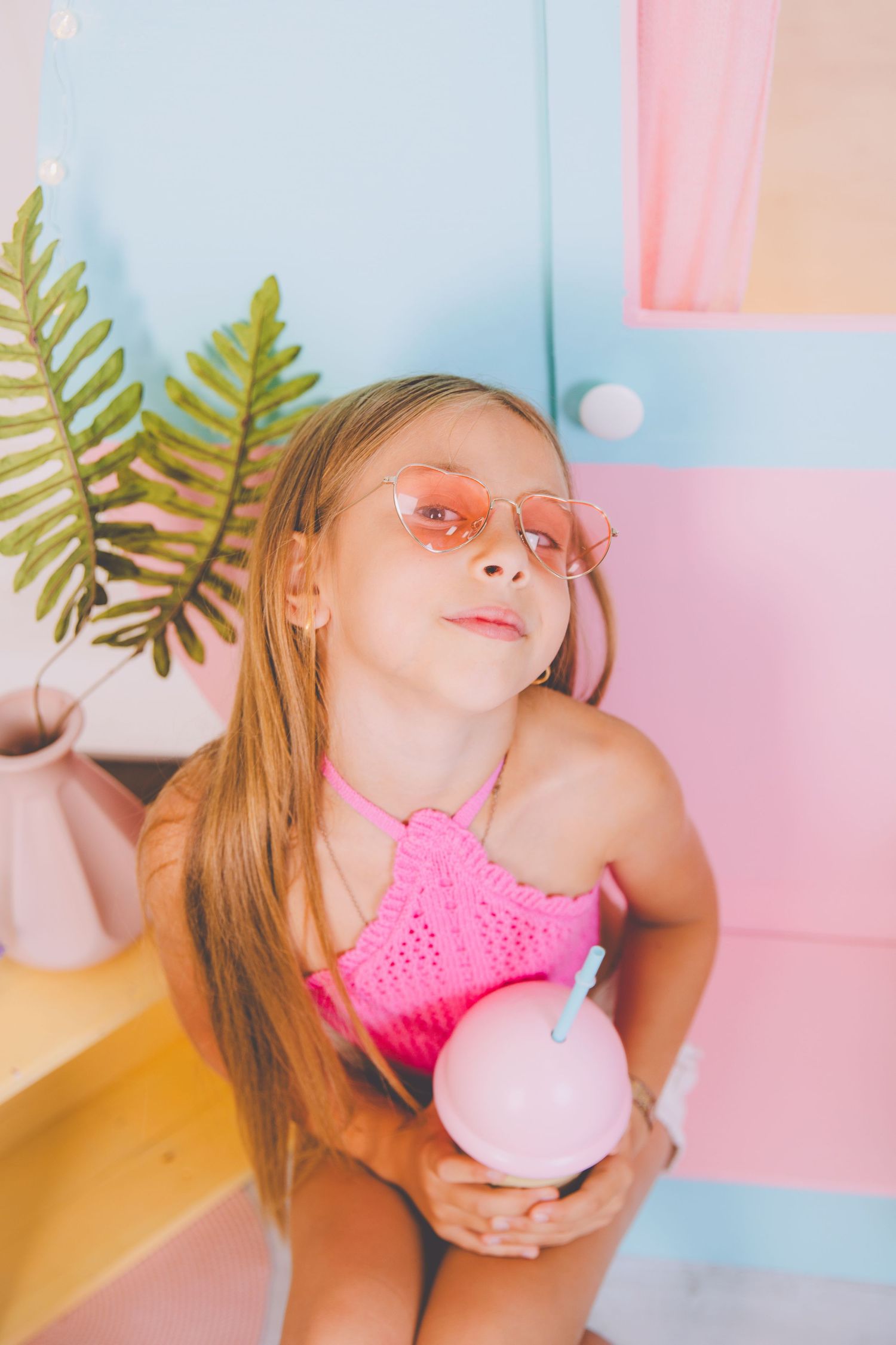 Una niña con traje de baño rosa y gafas de sol en forma de corazón sostiene una bebida rosa durante su sesión de fotos.