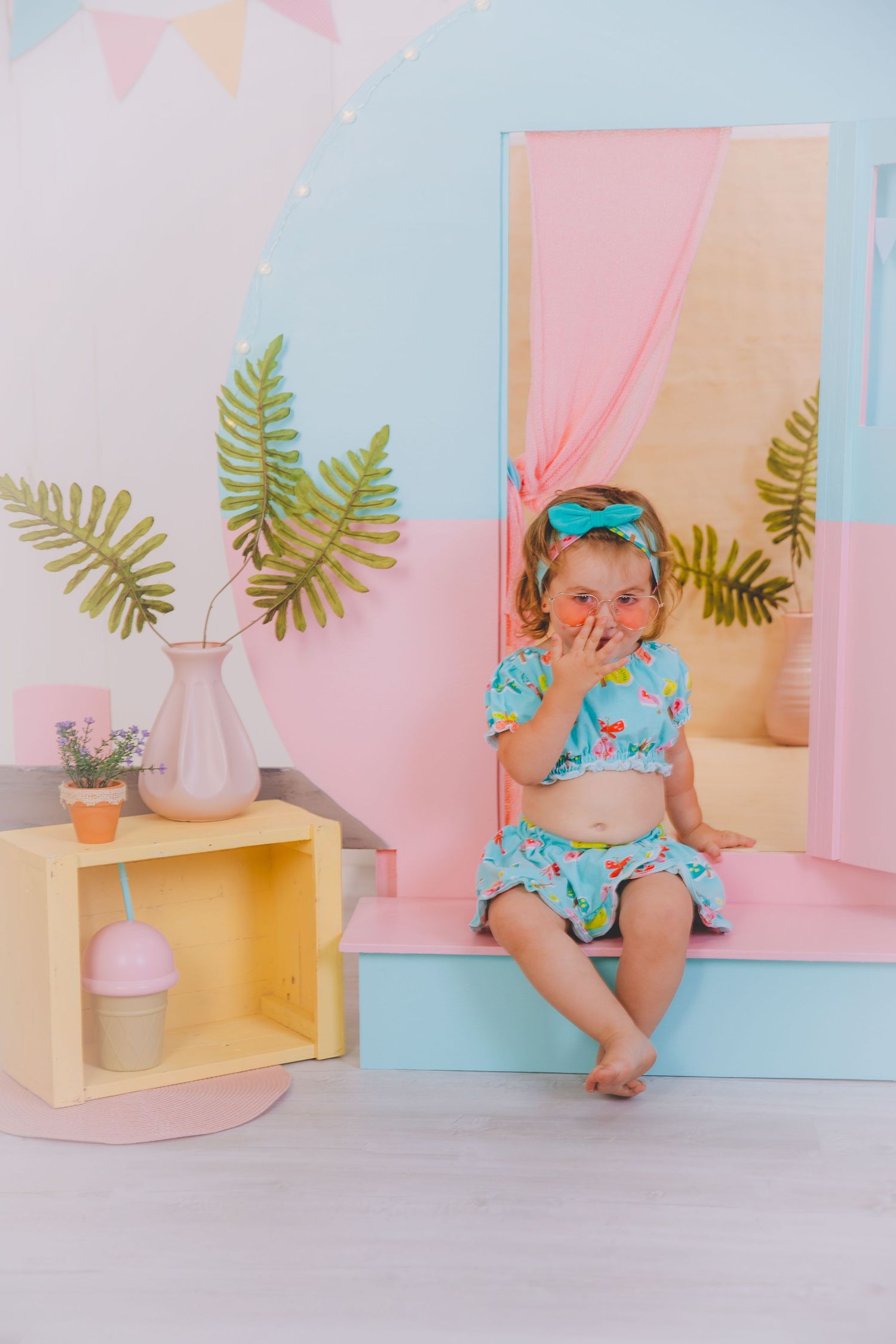 Una niña pequeña con ropa turquesa y una diadema con lazo está sentada posando para su sesión de fotos en el decorado de verano.