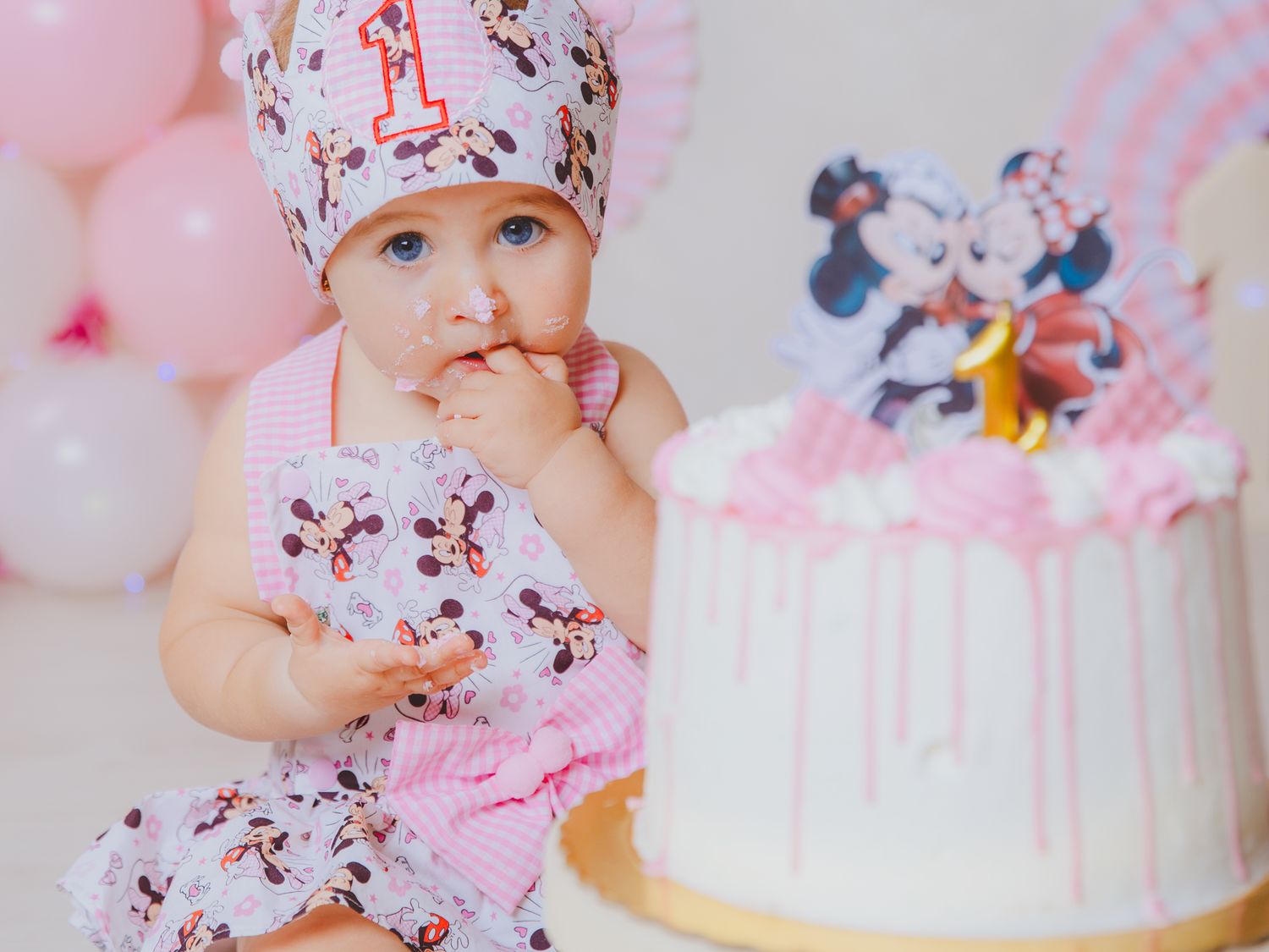 Bebé vestida con un traje de ratoncita con lazo está sentada junto a una tarta blanca y rosa con glaseado para celebrar su primer cumpleaños