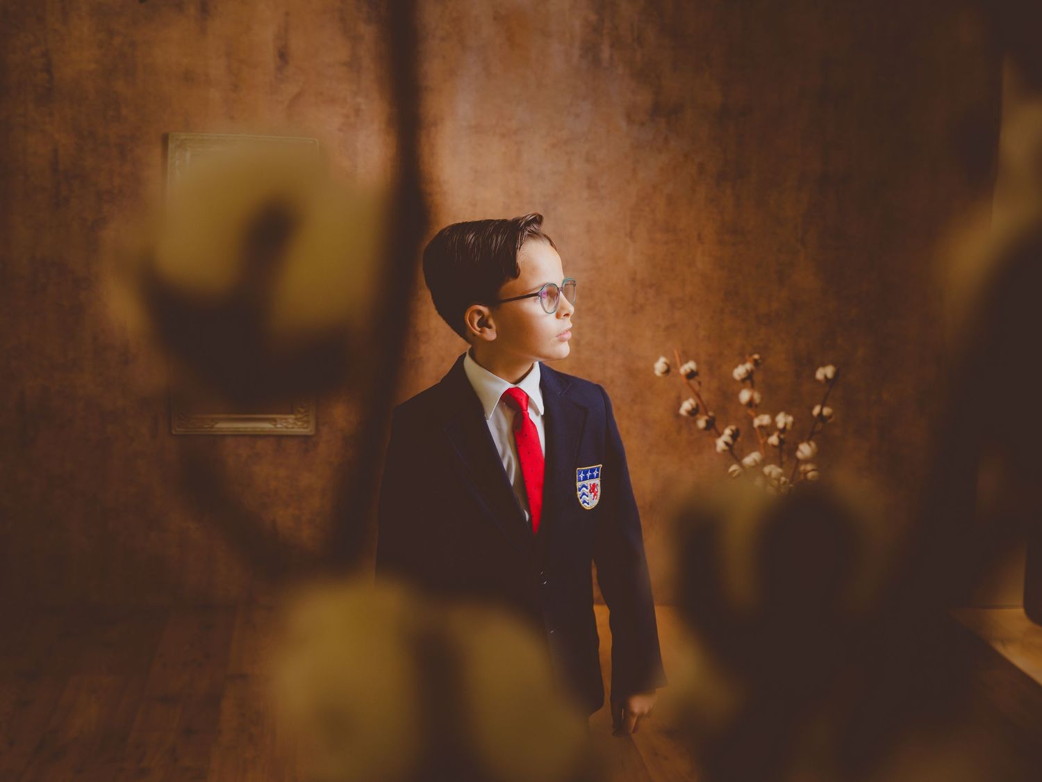 Niño con uniforme escolar de comunión y corbata roja de pie junto a flores secas sobre un fondo de madera.