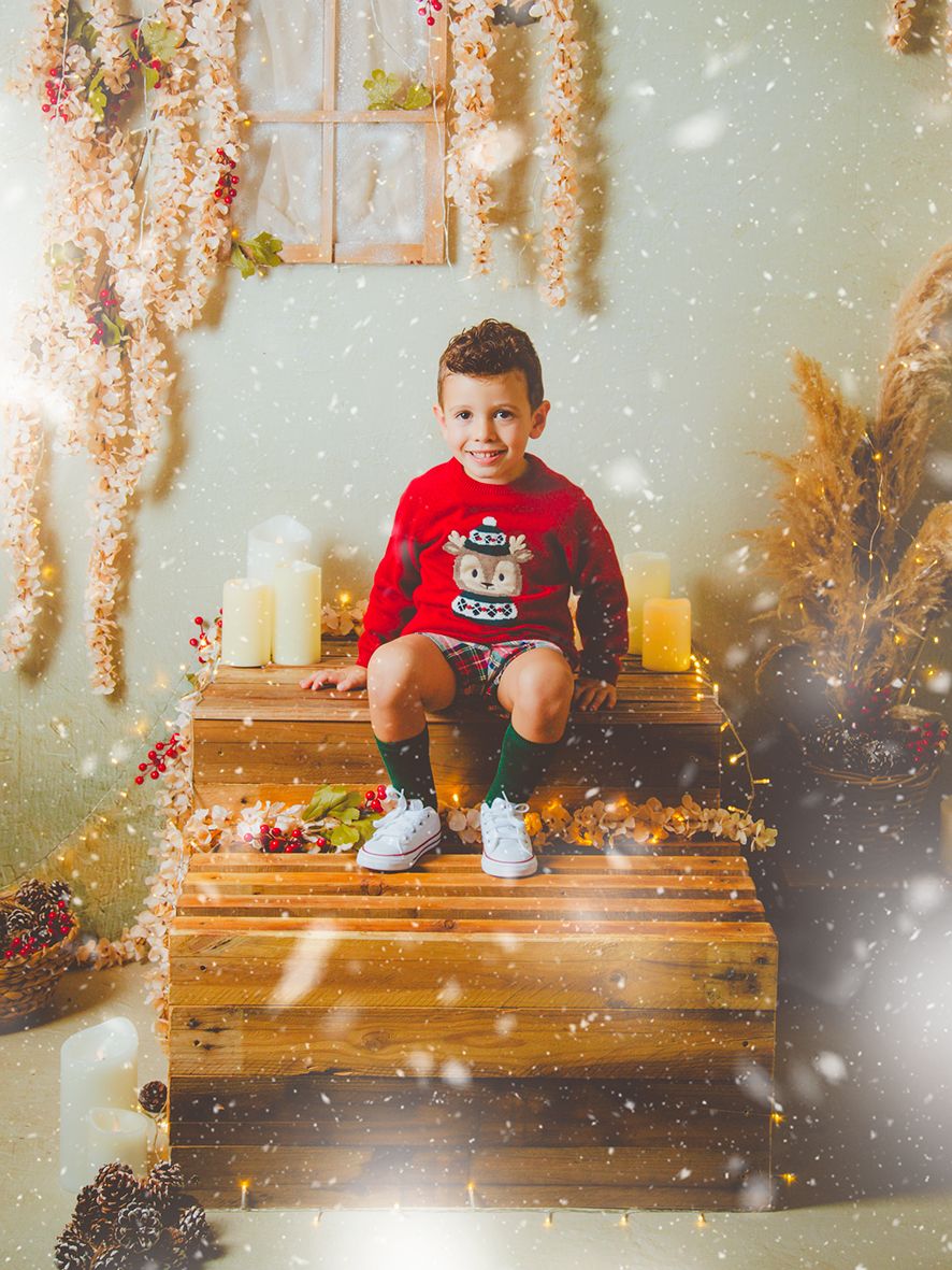 Niño sonriendo con un jersey rojo navideño está sentado sobre una escalera de madera rodeado de velas y adornos navideños en su sesión.