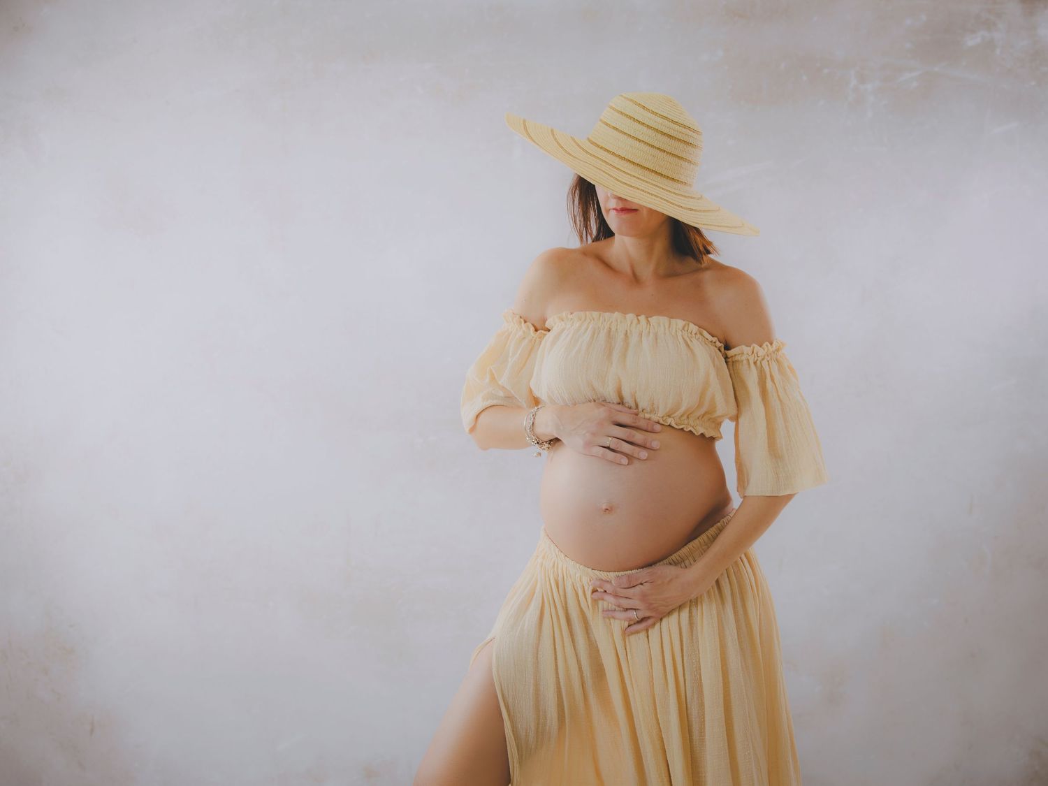 Una mujer embarazada con un vestido amarillo y un sombrero acaricia su barriga en su sesión de estudio.