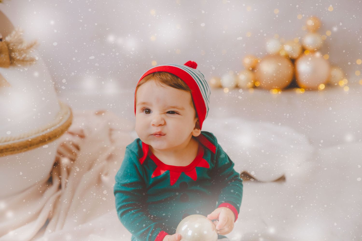 Adorable bebé vestido de elfo sosteniendo un adorno navideño con decoraciones doradas y un fondo de Navidad.