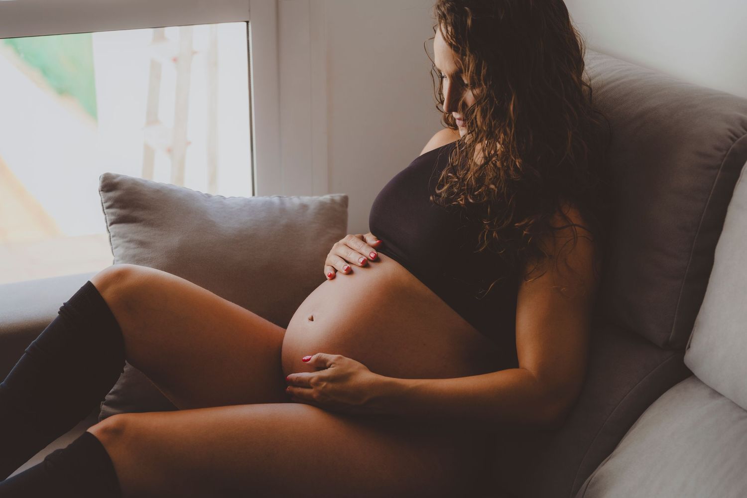 Fotografía de una embarazada sentada en un sofá, tocando su barriga durante su sesión fotográfica en casa.