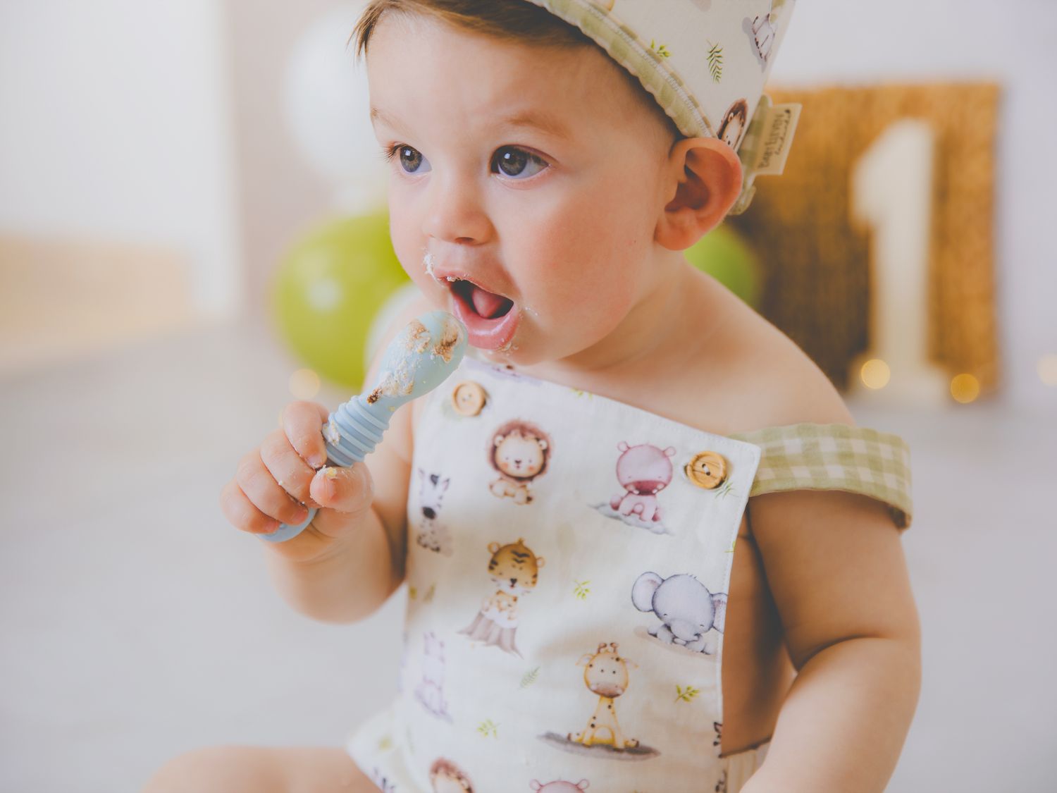 Precioso niño vestido con ranita de animales y cuadros vichy llevándose la cuchara de tarta de su sesión de cumpleaños a la boca.