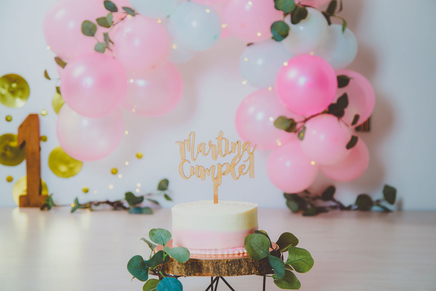Fotografía de una tarta de primer cumpleaños en un decorado muy elegante en tonos blanco, rosa dorado y detalles de hojas verdes.