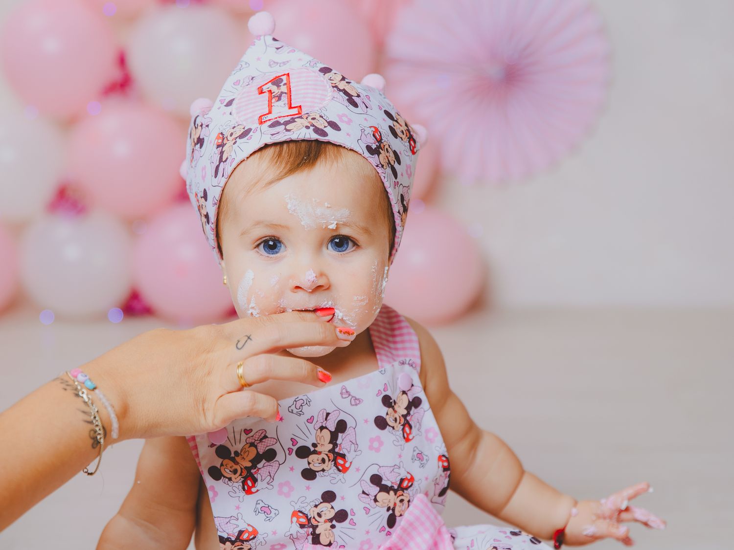 Una bebé con un vestido de ratoncita con lazo y corona en la cabeza en una sesión de fotos de cumpleaños, come tarta del dedo de mamá.