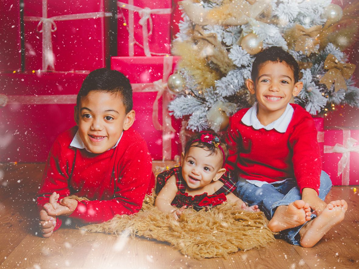Tres hermanos con conjuntos rojos navideños sonríen juntos junto a un árbol decorado con regalos rosas.