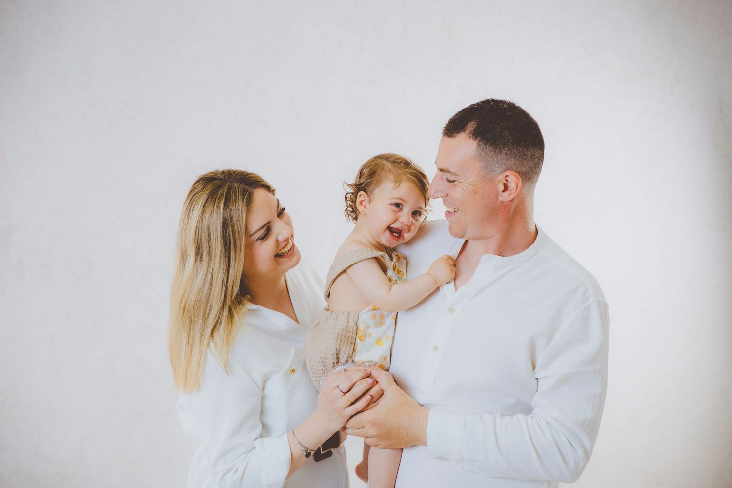 Familia feliz con su niña pequeña sonriente en brazos de su madre y de su padre, ambos vestidos de blanco.