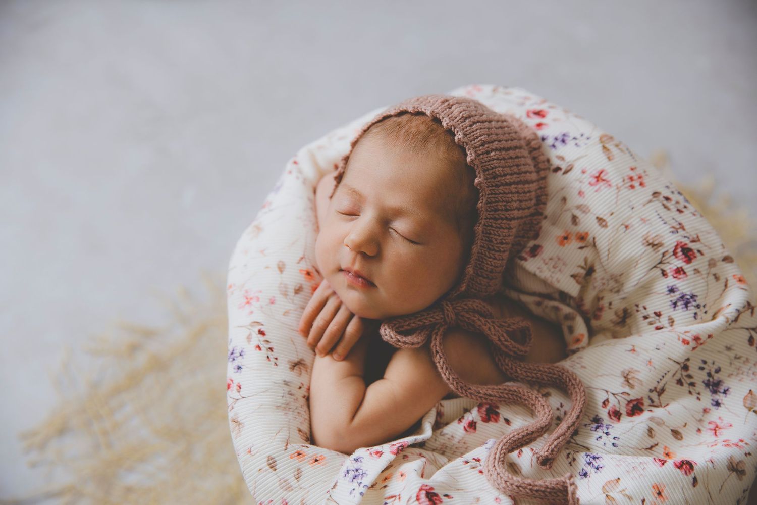 Un bebé recién nacida duerme plácidamente en un cuenco con tela de flores, con un suave gorrito de punto y las manos bajo la barbilla.