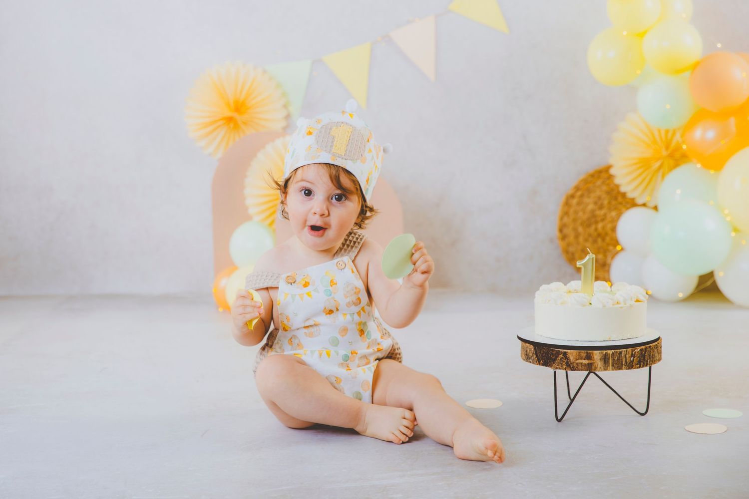 Una bebé con gesto sorprendido y corona de cumpleaños junto a una tarta y decoración con globos tonos pastel.