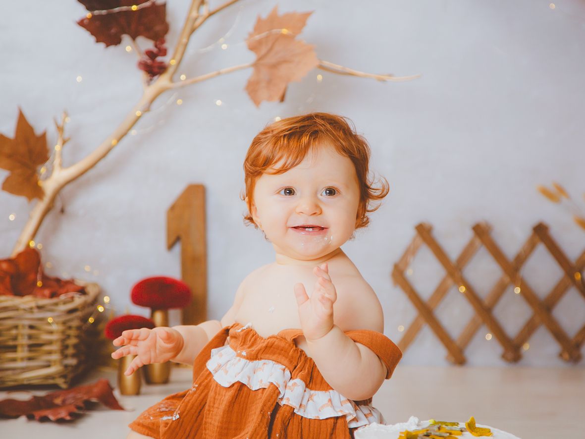 Una alegre bebé pelirroja con un vestido color naranjado saluda durante la sesión de fotos de su primer cumpleaños con temática otoñal.
