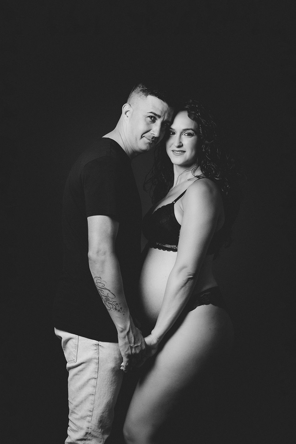 Fotografía en blanco y negro de una pareja mirando a cámara y agarrados de la mano en su sesión de embarazo.