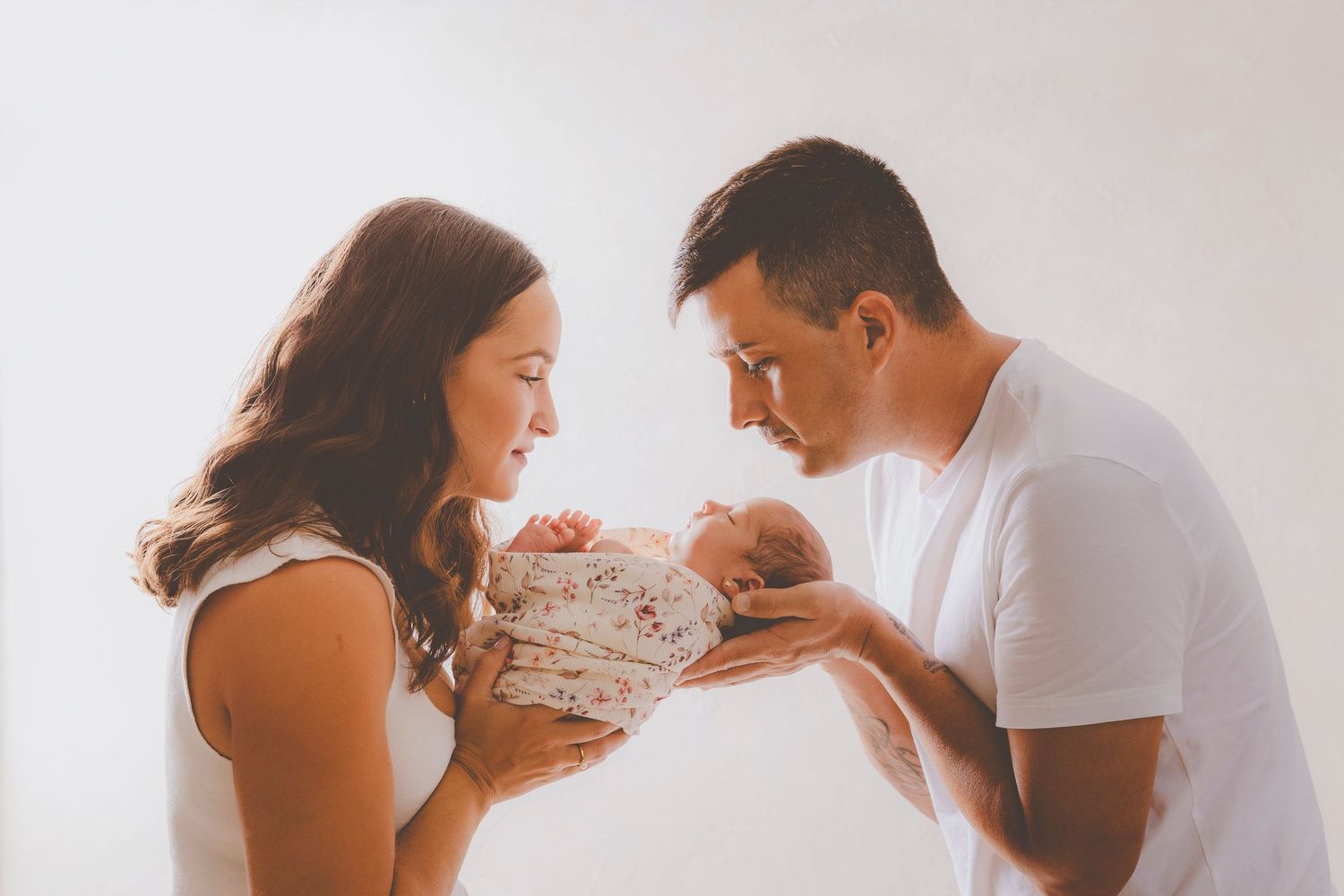 Una pareja mirando con ternura a su bebé recién nacido, sostenido entre ellos, sobre un fondo blanco, en su sesión de newborn.