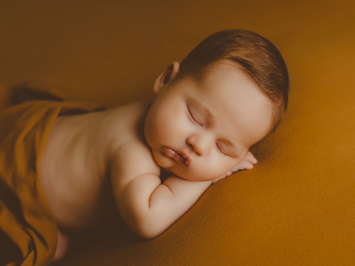 Retrato de estudio de un bebé recién nacido durmiendo sobre una manta de color mostaza con las manos metidas bajo la barbilla.