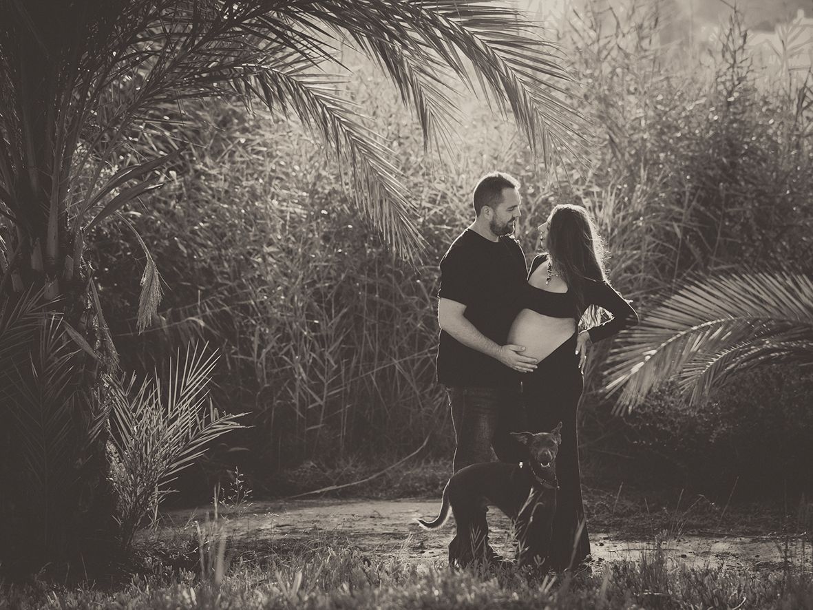 Fotografía en blanco y negro de estilo vintage de una pareja abrazándose entre palmeras con su perro.