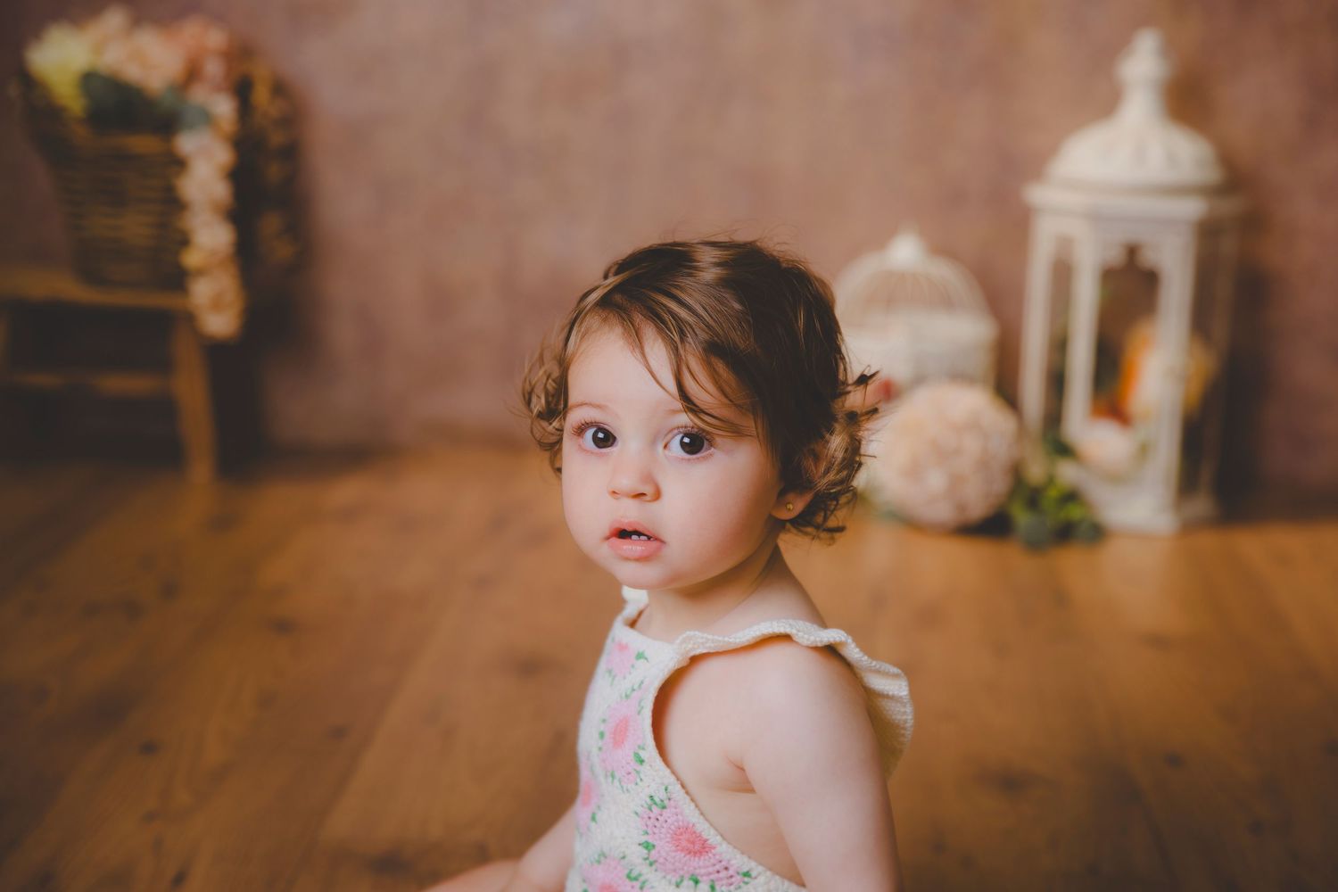 Retrato de una niña pequeña con un vestido de flores está sentada en un suelo de madera rodeada de faroles y flores.