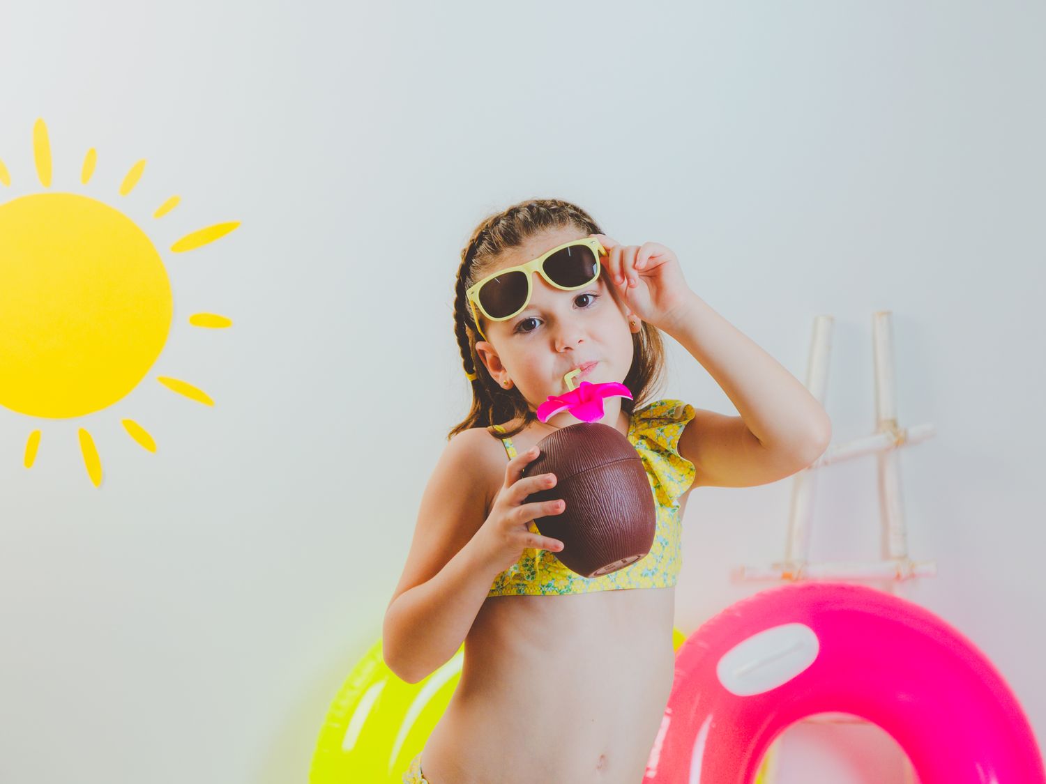 Niña en bikini con gafas de sol bebiendo de un coco con coloridos flotadores de piscina y el sol de fondo en su reportaje de verano.