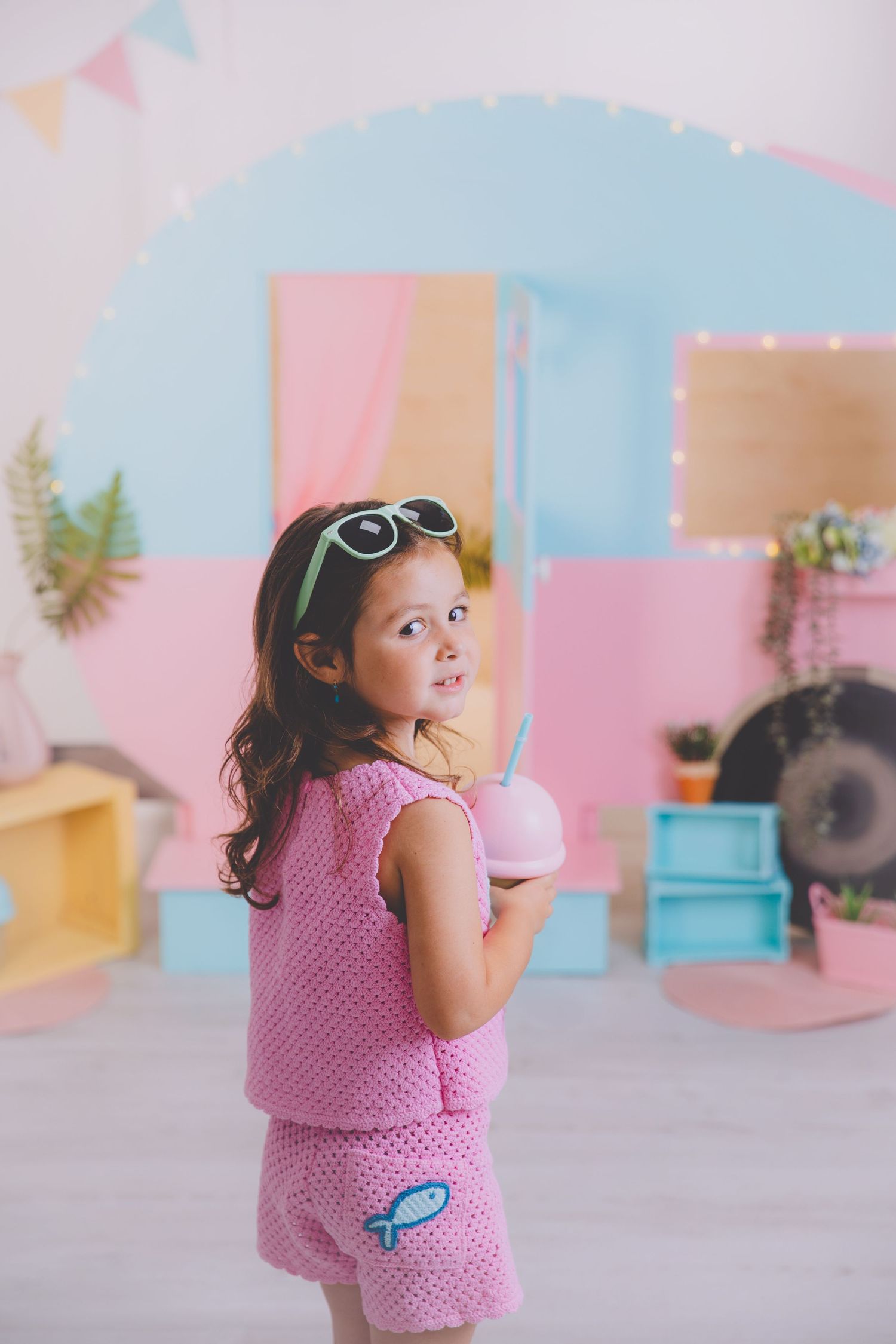 Una niña pequeña vestida de rosa sostiene un vaso en forma de helado rosa mientras posa frente a un colorido fondo de estudio de verano.