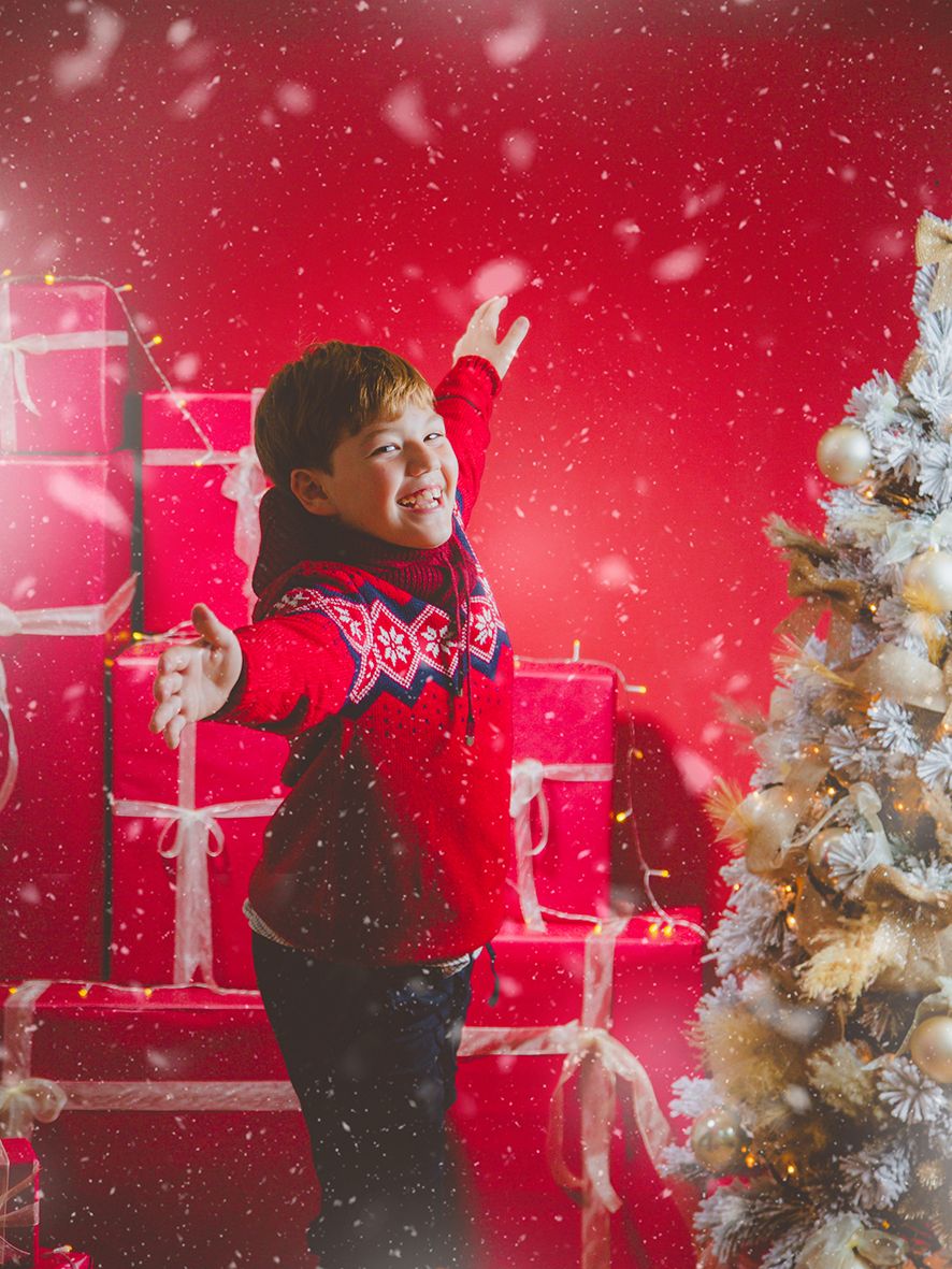 Niño alegre con jersey nórdico rojo intenta atrapar copos de nieve junto a un árbol de Navidad con regalos.