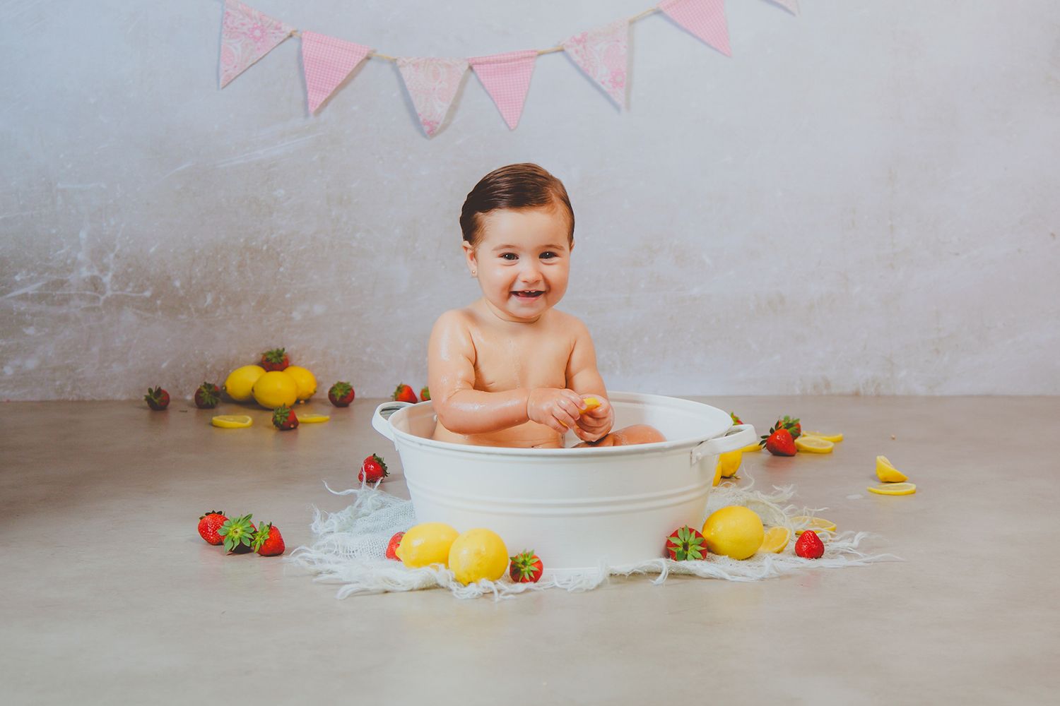 Bebé feliz riendo en una bañera blanca rodeada de fresas y limones en el suelo durante su sesión de cumpleaños.