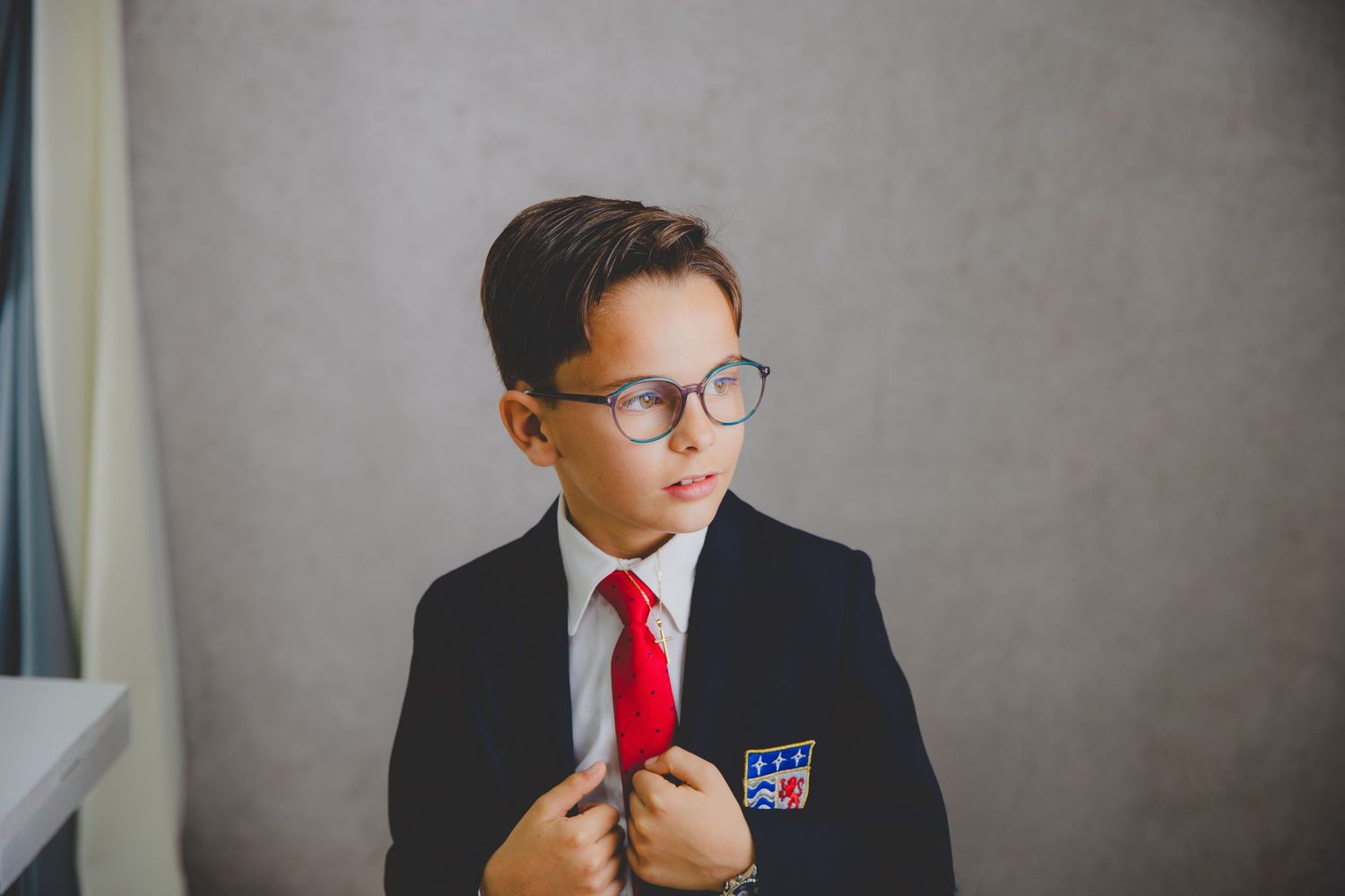 Un niño con gafas se sujeta la chaqueta de su traje de comunión con corbata roja en un fondo de estudio gris.