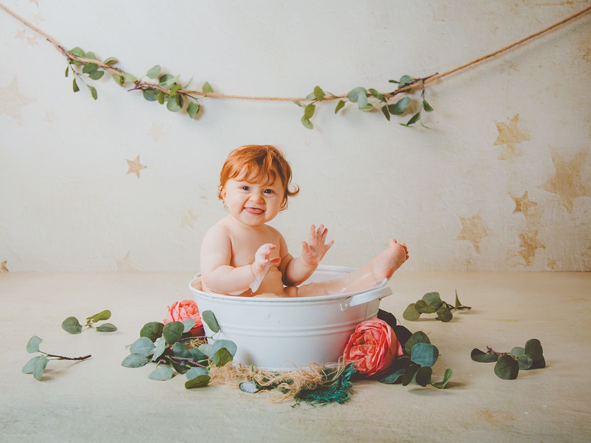 Una bebé pelirroja y feliz sonríe en una bañera blanca rodeado de rosas y eucalipto en un estudio con fondo de estrellas.