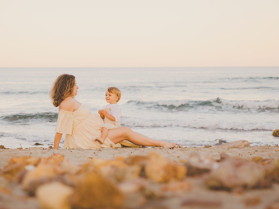 Imagen de una madre embarazada y su bebé sentados juntos en una playa al atardecer, junto a las olas del mar en Genoveses.