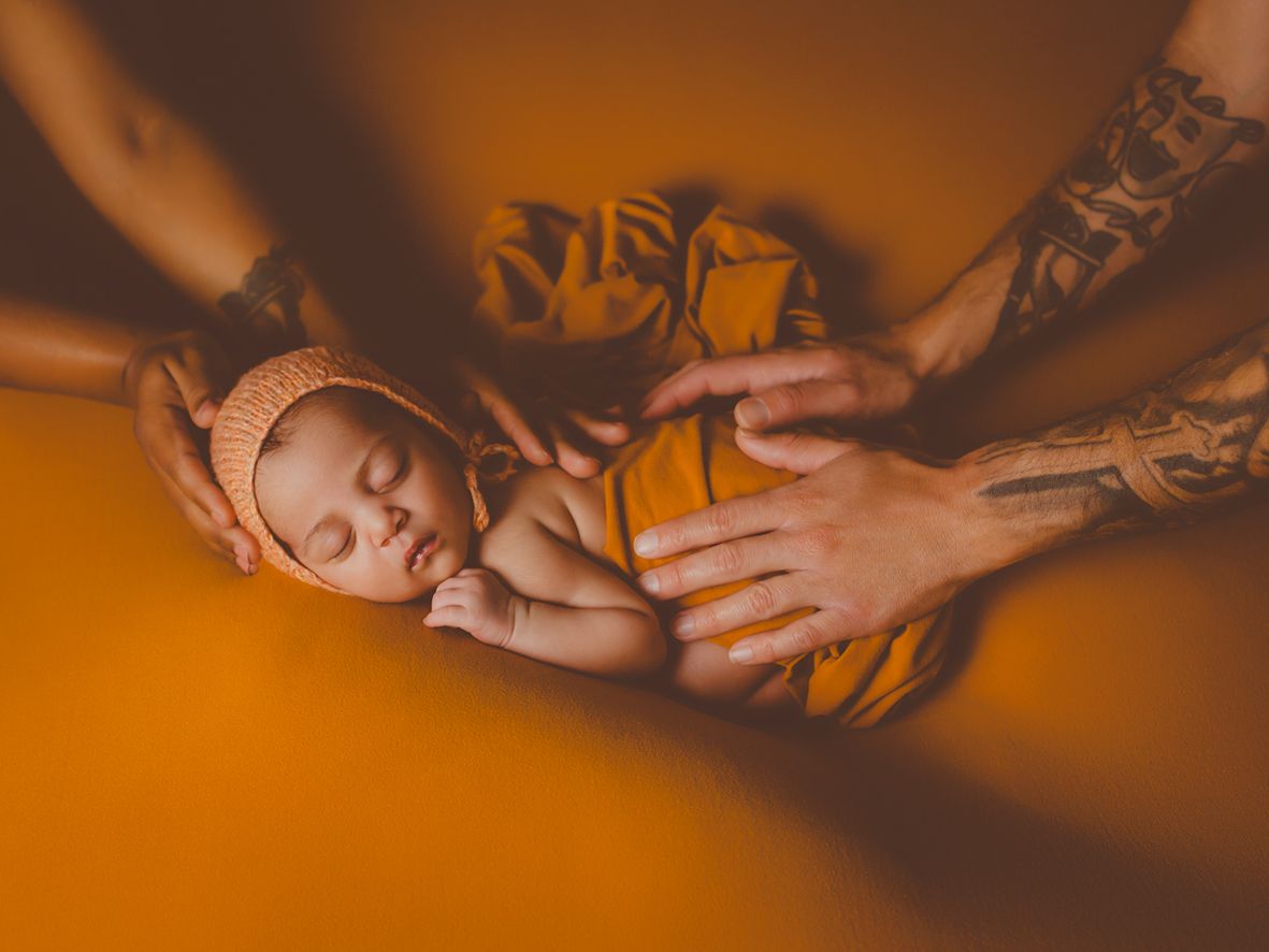 Padres tatuados sostienen con ternura a su bebé recién nacido dormido durante la sesión de fotos, envuelto en una manta de color naranja.