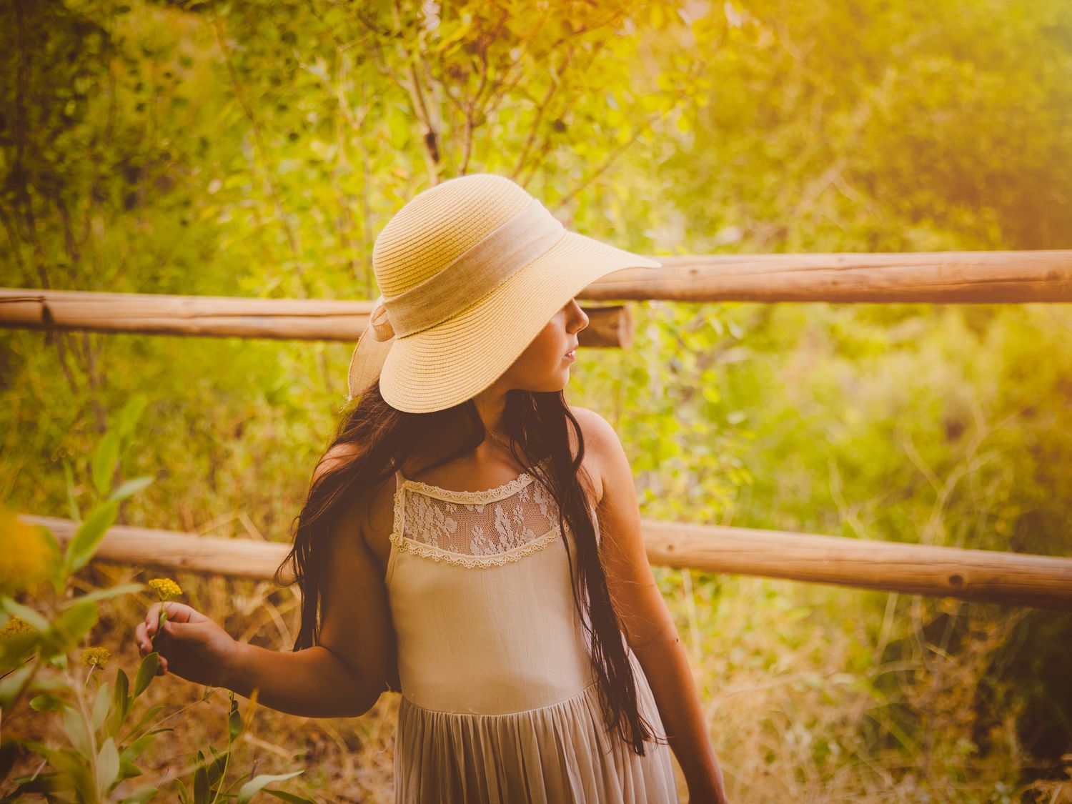 Foto de exteriores de comunión de una niña con sombrero de ala ancha y vestido crema junto a una valla de madera.