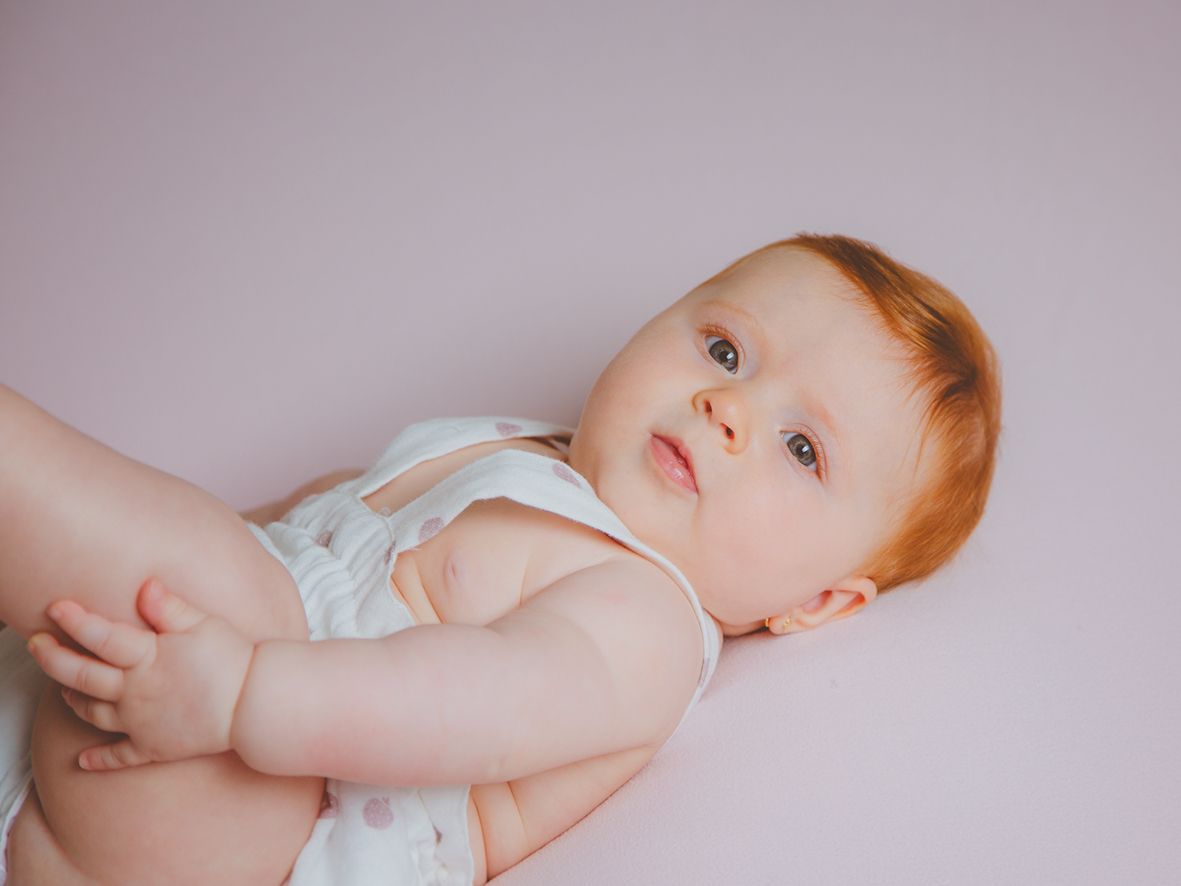 Fotografía de bebé pelirroja con un pelele blanco, tumbado sobre un fondo rosa con los pies en alto.
