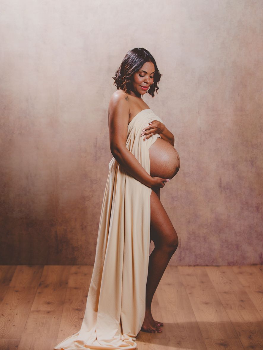 Elegante mujer embarazada envuelta en tela color crema posando en una sesión de fotos de maternidad profesional en estudio.