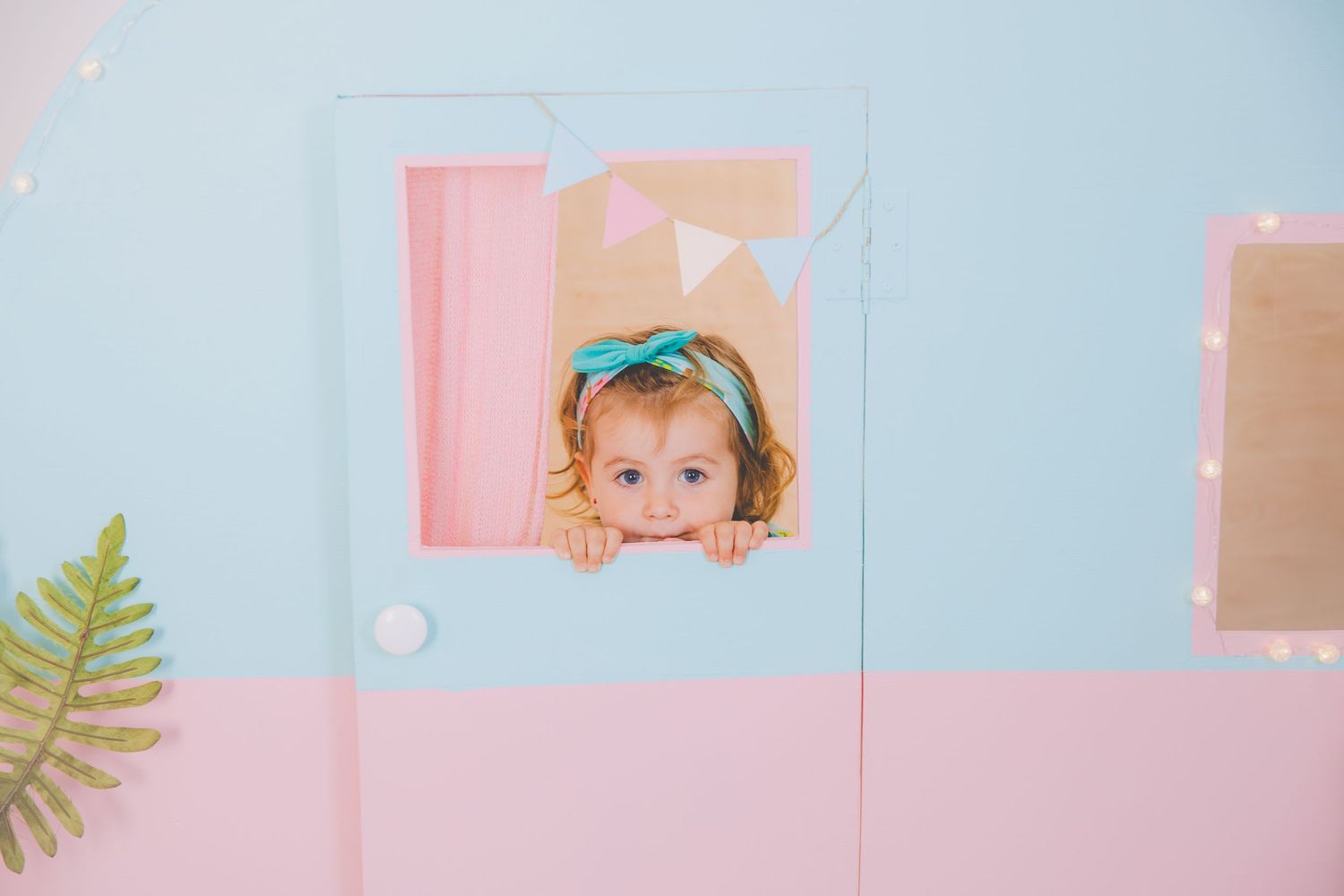 Una adorable niña pequeña con un lazo color turquesa se asoma por la ventana del decorado de caravana de la sesión de verano.