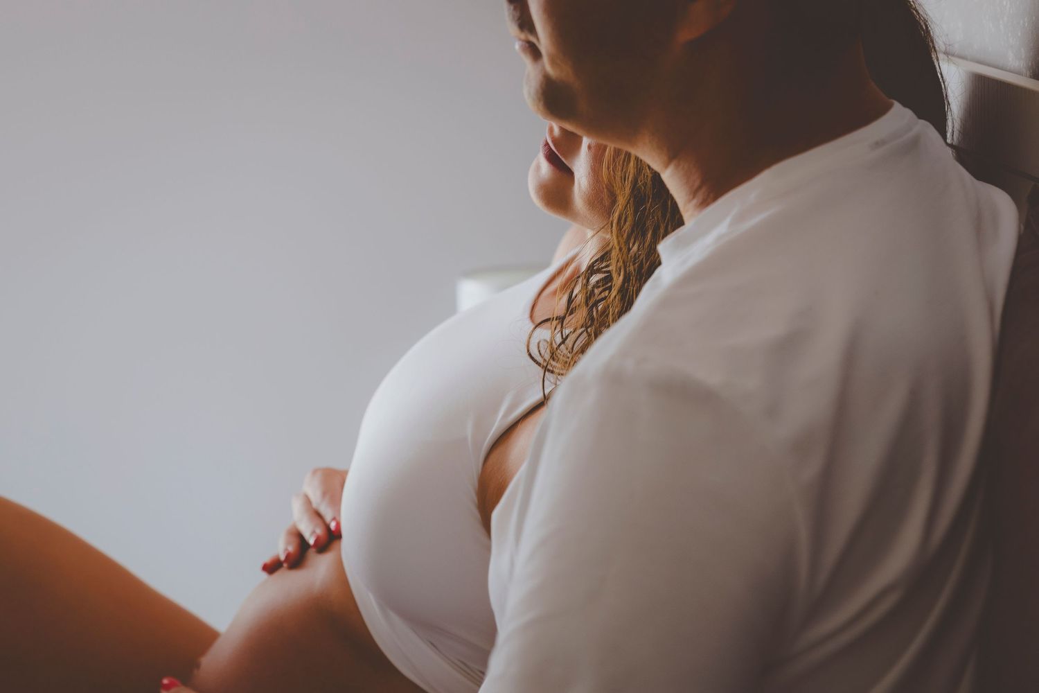 Una mujer embarazada, vestida con ropa blanca, acaricia su barriga cerca de una ventana luminosa.