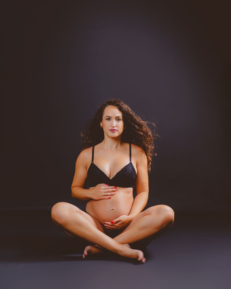 Mujer embarazada sentada con las piernas cruzadas, vestida con lencería negra, con un fondo oscuro de estudio en su sesión de embarazada.