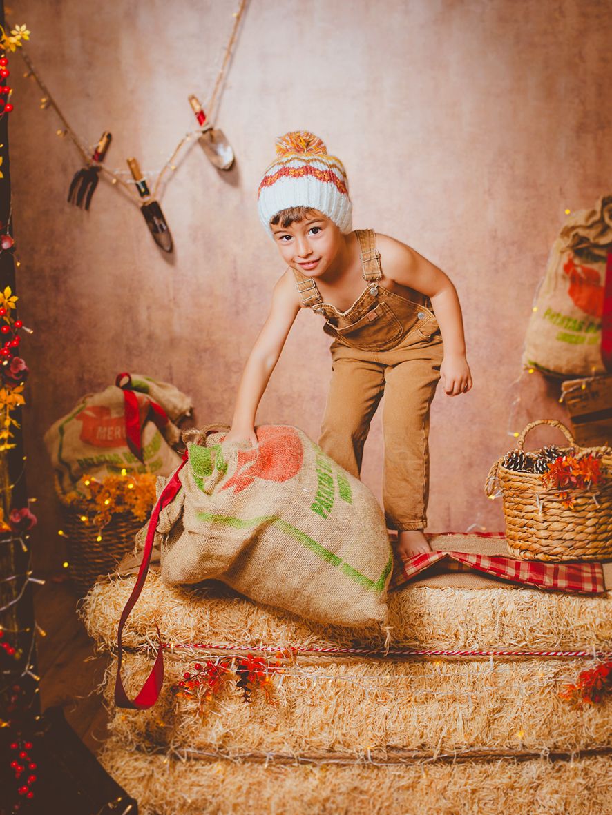 Fotografía de niño con peto y gorro de invierno levanta un saco de arpillera sobre fardos de heno en una escena rústica de Navidad.