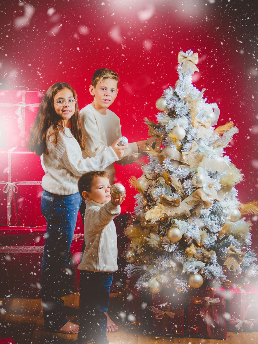 Tres niños decorando un árbol de Navidad blanco y dorado con regalos envueltos sobre un fondo de estudio rojo nevado.