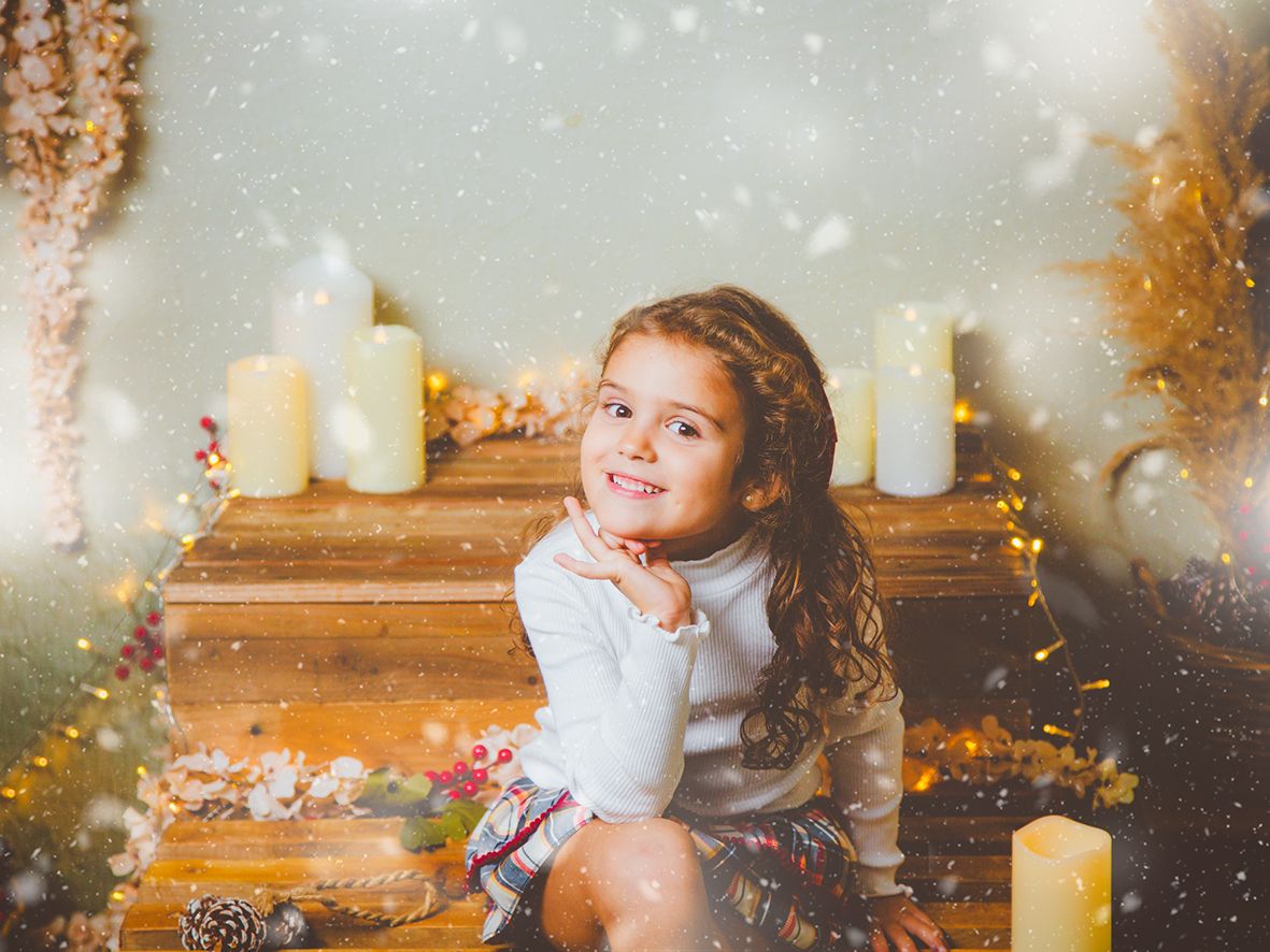 Una niña posando en su sesión de Navidad sonriente sentada en un decorado de madera rodeada de velas y nieve.