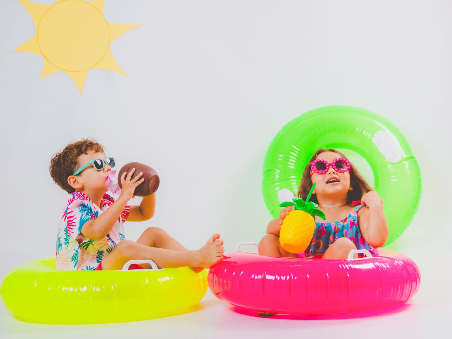 Dos niños están sentados en coloridos flotadores sosteniendo fruta en su divertida sesión de verano.